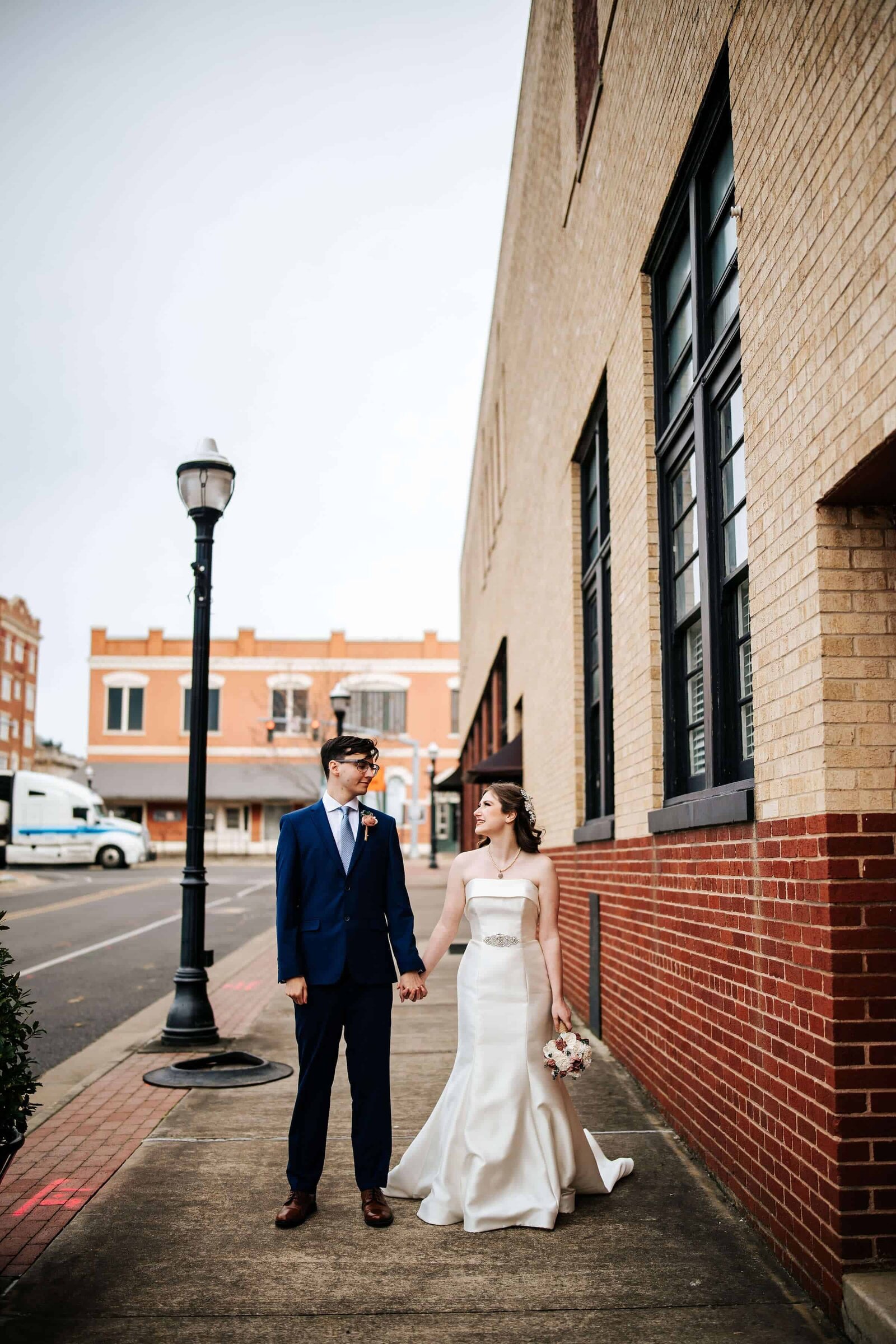 A bride in a white dress and a groom in a blue suit hold hands while walking on a city sidewalk in Ruston, Louisiana next to a brick building, looking at each other lovingly.