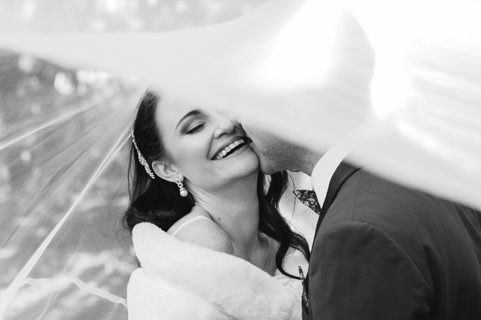 Candid moment of bride smiling and closing her eyes while groom kisses her cheek, veil gently covering them.