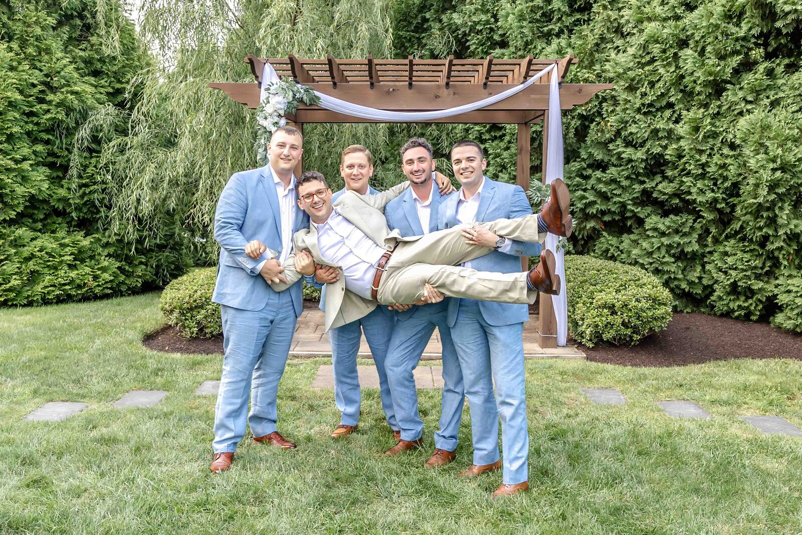 The groom is playfully lifted by his groomsmen in front of a decorated wooden ceremony arbor, surrounded by lush greenery, captured during a Wilmington, DE wedding, showcasing fun and lighthearted moments.