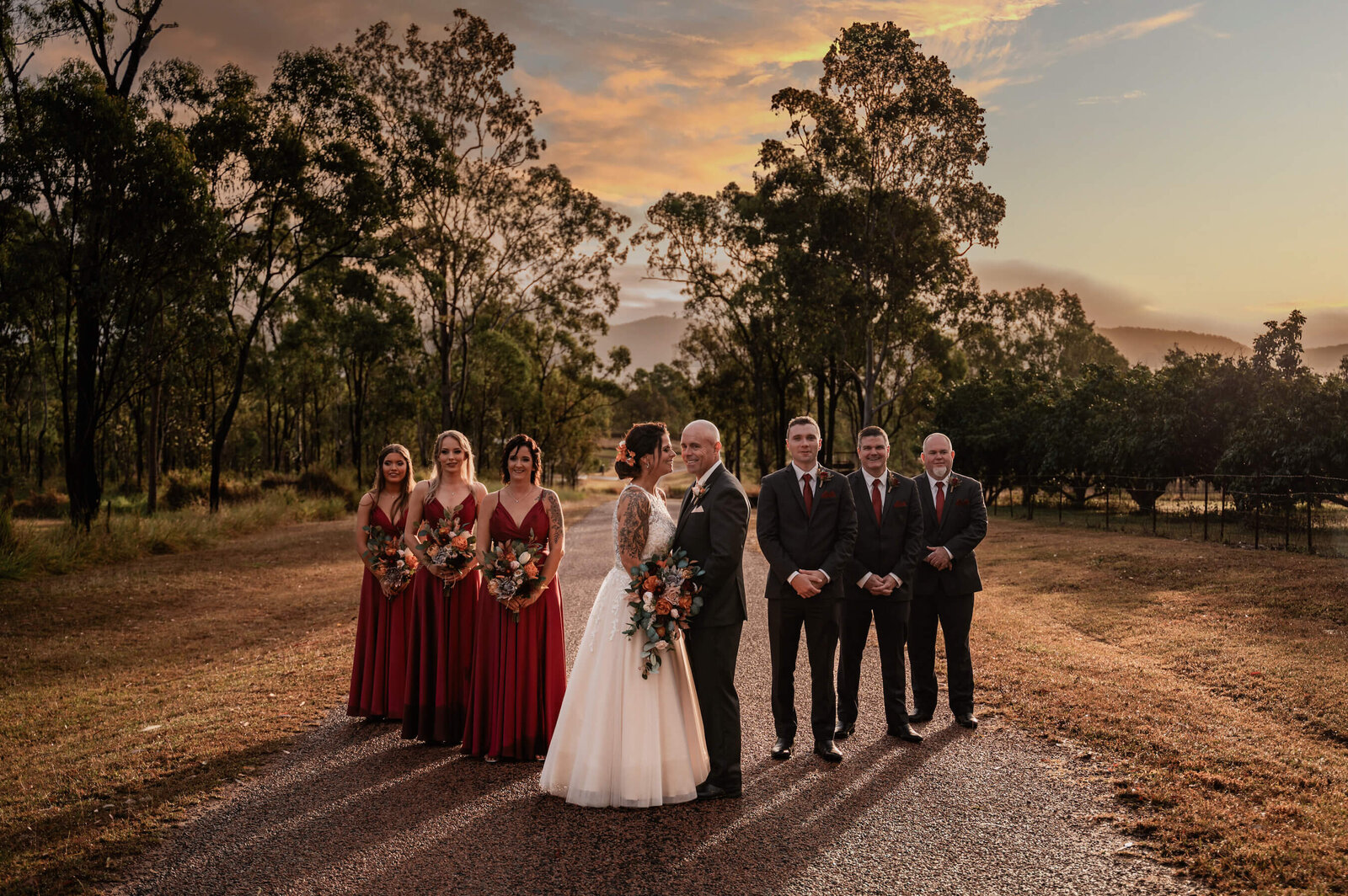 A wedding party stands together for a photo, illuminated by the warm glow of a sunset in the background.