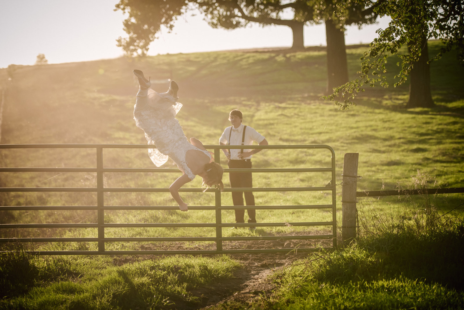 bride jumping over a fence in her wedding dress while groom watches