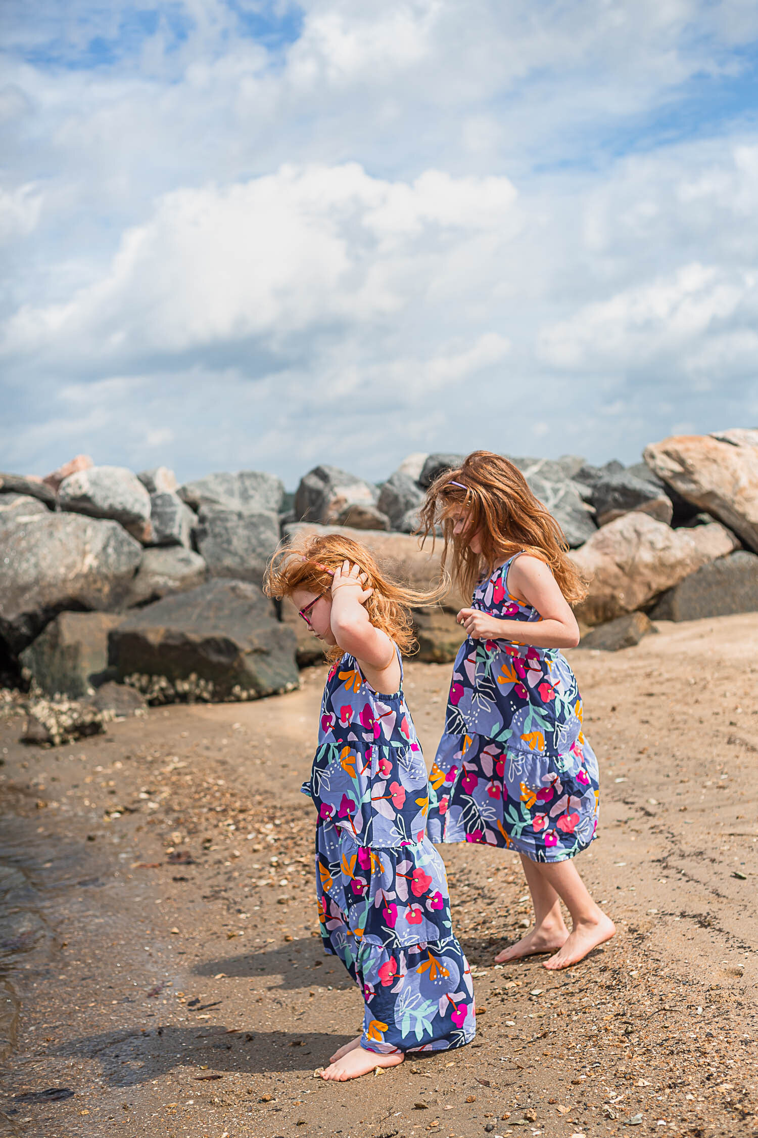 beach family portraits with two sisters in matching dresses and red hair walk up to the shortline as the wind blows through their hair taken by best Newport News family photographers