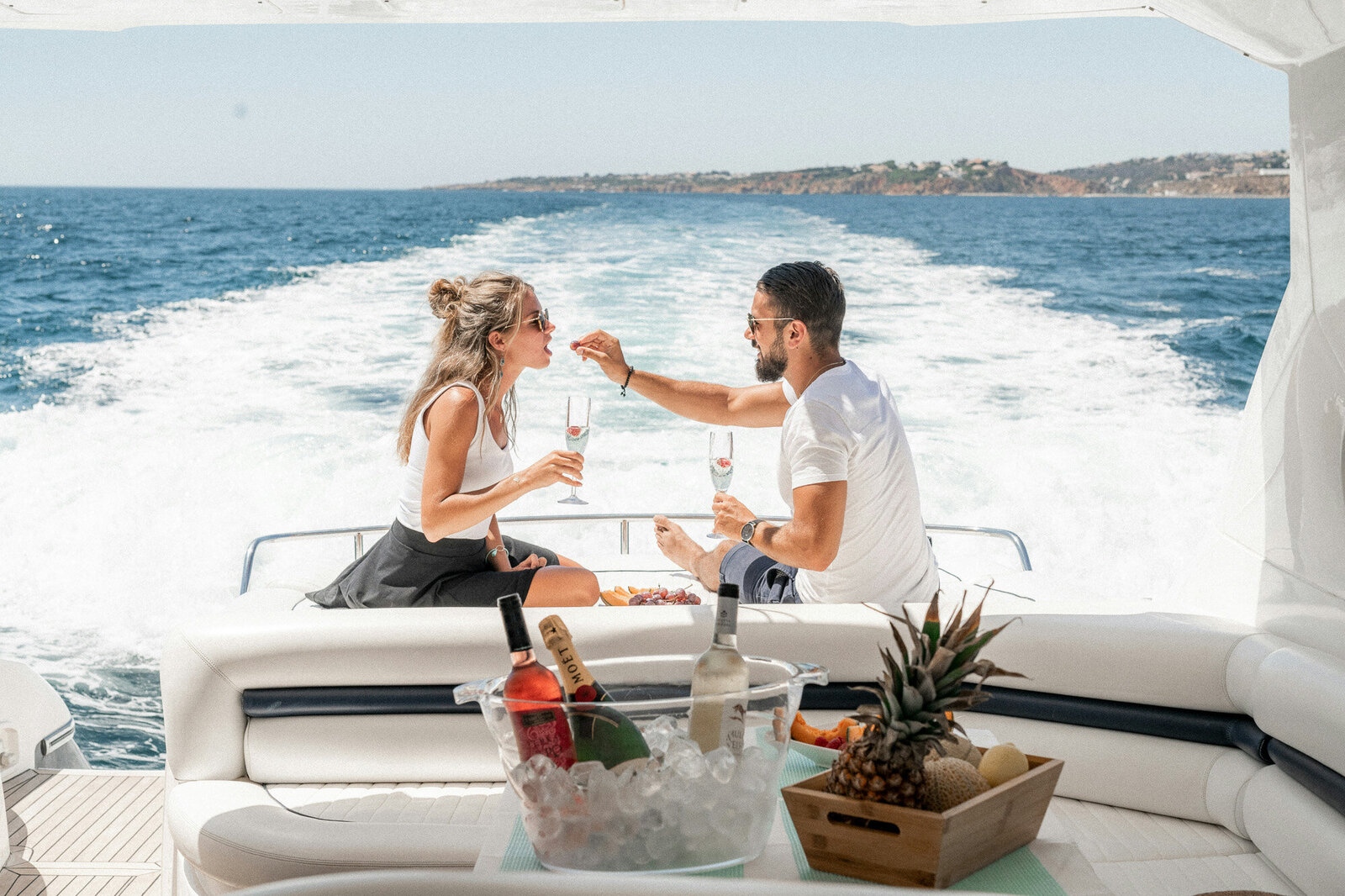 A couple sits on a yacht's deck, toasting with champagne glasses, man feeds the woman, while a bucket of ice with drinks and a tray holding a pineapple and banana are in the foreground