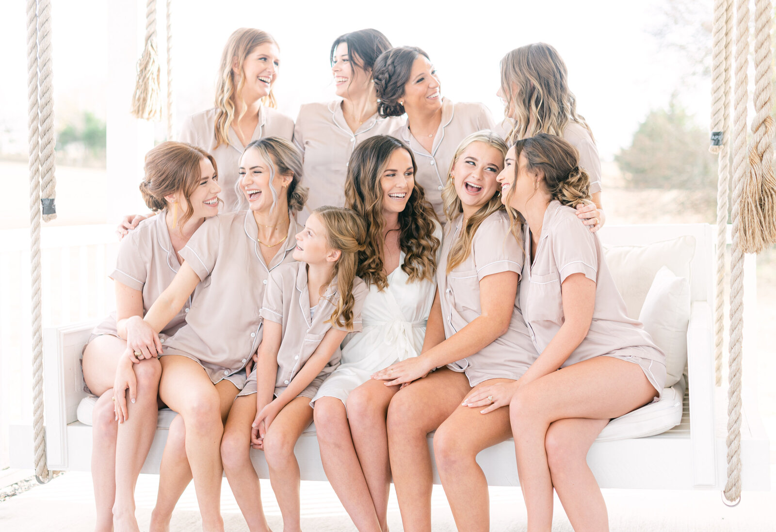 Portrait of a group of bridesmaids wearing cream-colored loungewear sitting on a daybed with a bride in a silk robe outdoors.