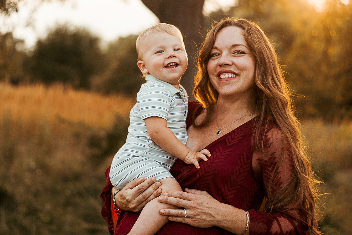 Mother with Toddler on arm. Standing in a meadow at sunset, both smiling. Mommy and me photo.  Family Photography.