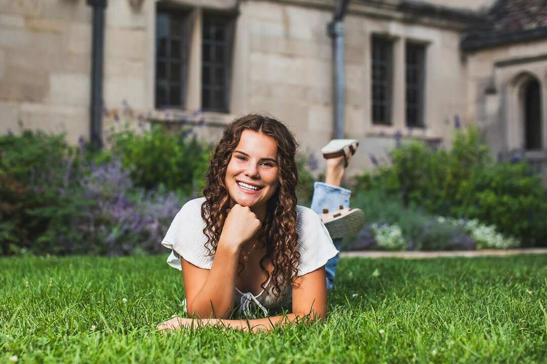 This athletic curly haired senior girl poses on her belly for her senior photo shoot in the grass near the mansion.