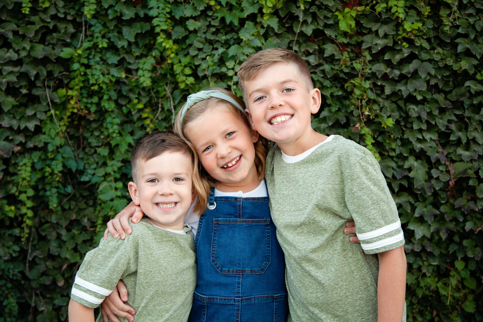 Close up of three siblings smiling in front of greenery.