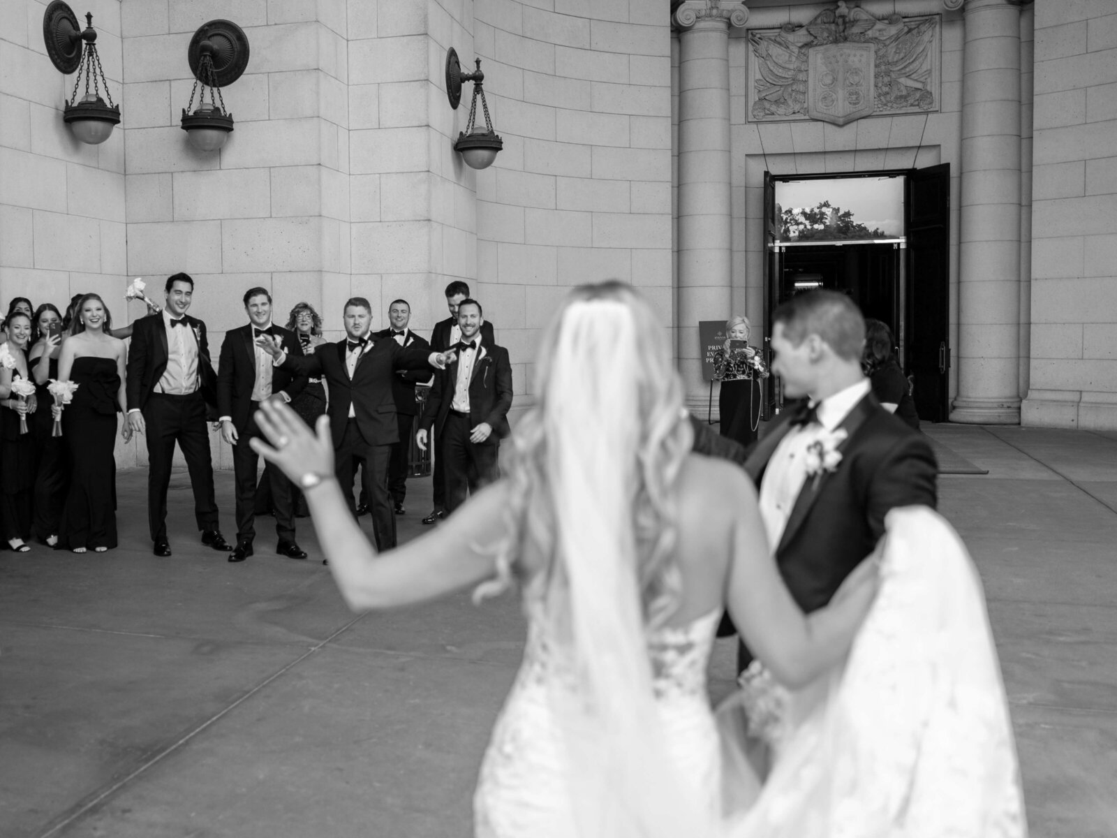 A bride and groom dance outside a building with open doors. The bride has long, wavy hair and wears a veil and gown, while the groom is in a suit. A group of guests stands nearby, clapping and smiling. The scene is in black and white.