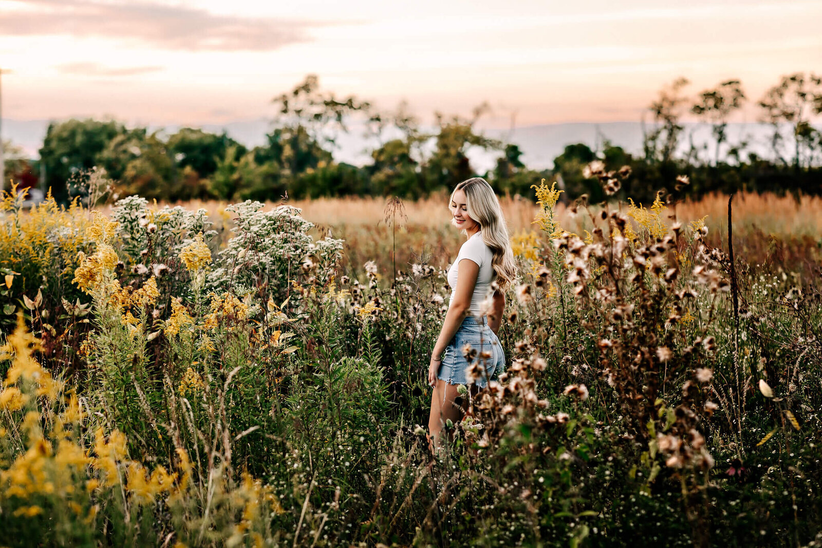 A person with long hair wearing a white top and denim shorts stands in a field filled with tall wildflowers and grass. The sky is painted with warm colors of a setting sun. Trees line the background.