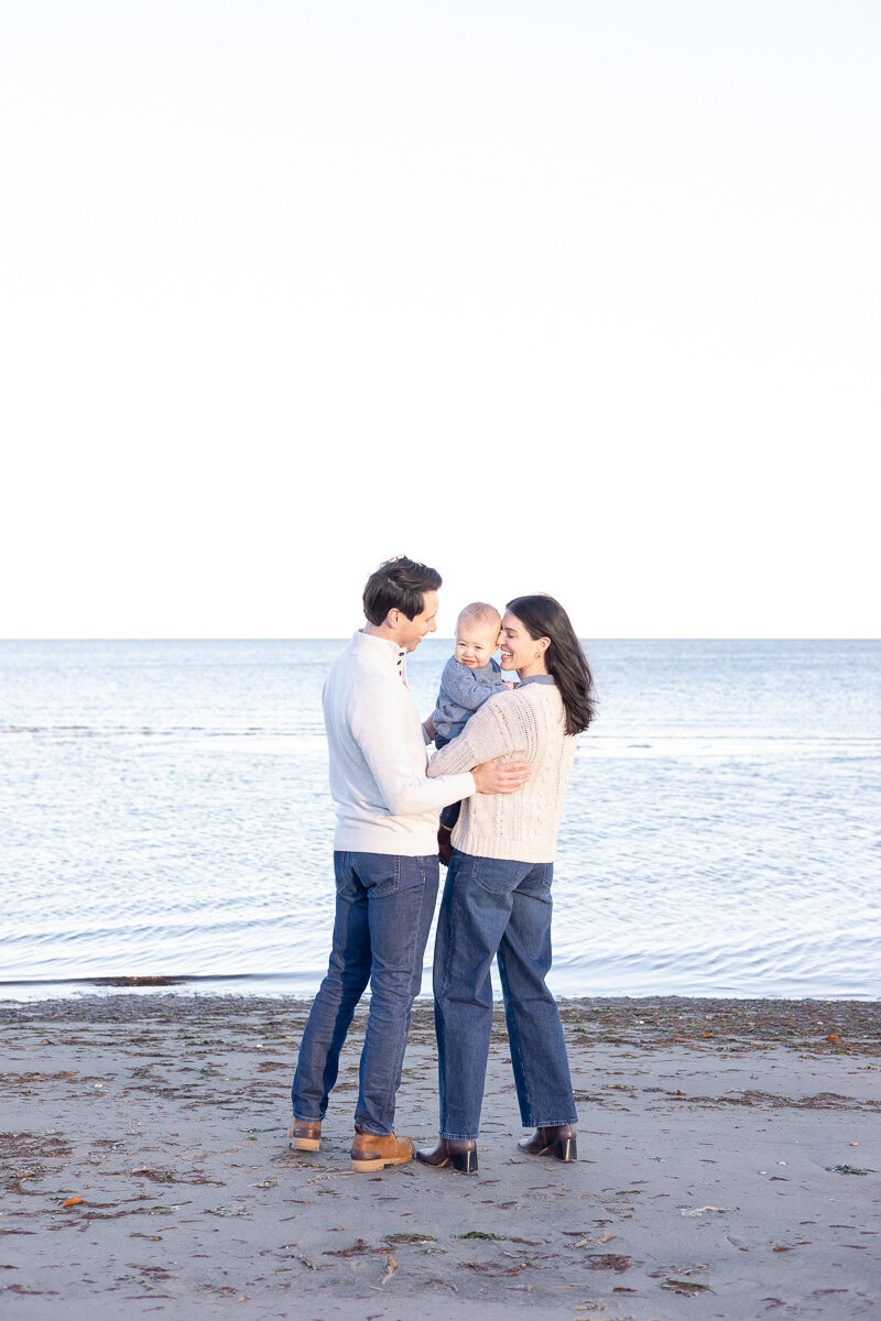 Young family smiling together during a candid beach photo session in Connecticut. Natural light family portraits by Nicole, a Fairfield County lifestyle photographer for laid-back, loving families.