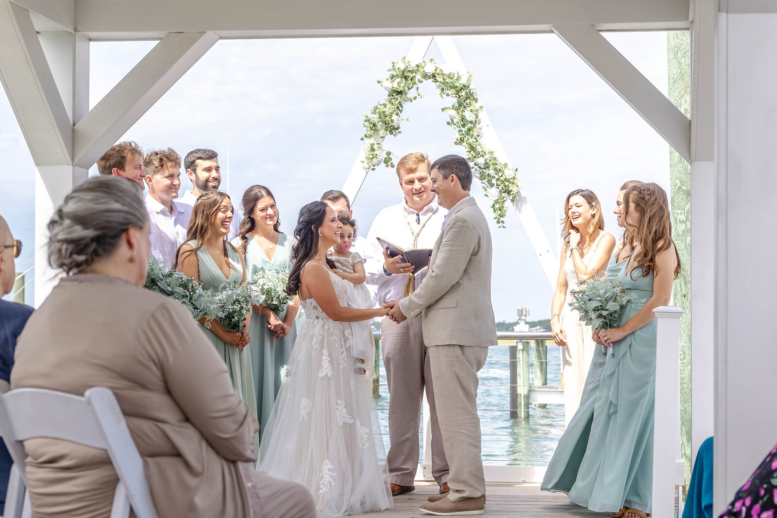 The bride and groom exchange vows under a white arbor adorned with greenery, surrounded by their bridal party and family, captured during a waterfront wedding in Wilmington, NC, blending elegance and natural beauty.