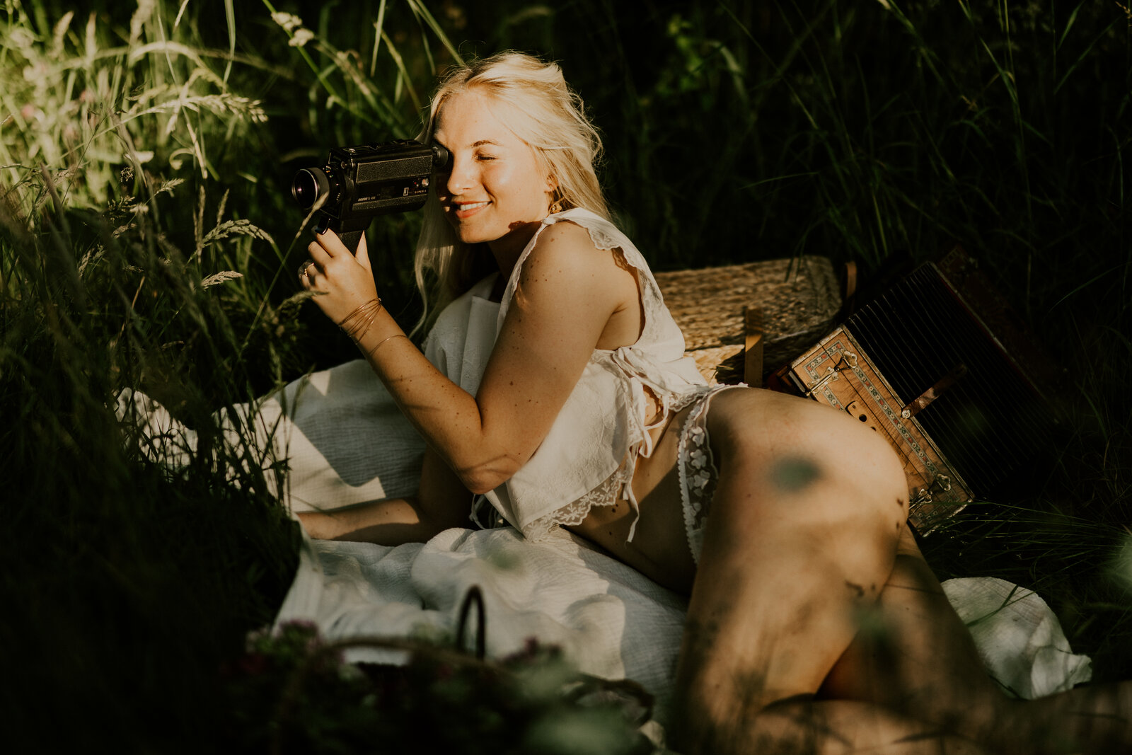Petite fille assise sur un drap et entourée de coussins à la plage, portant une robe blanche et des lunettes de soleil rondes, sourire espiègle. Photographie prise par Laura, photographe en vendée.