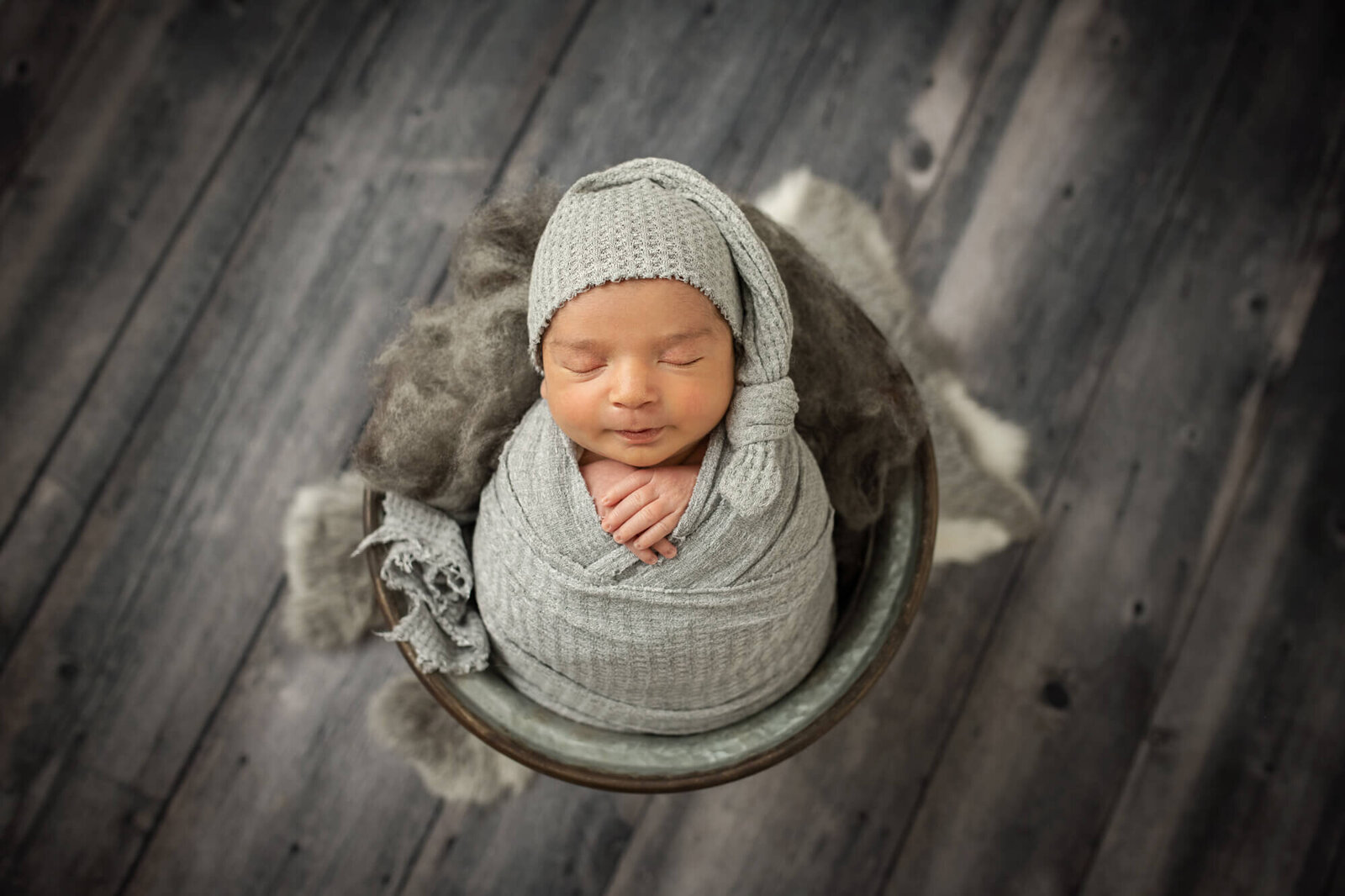 Sleeping baby boy in bucket with grey wood floor wearing sleepy cap
