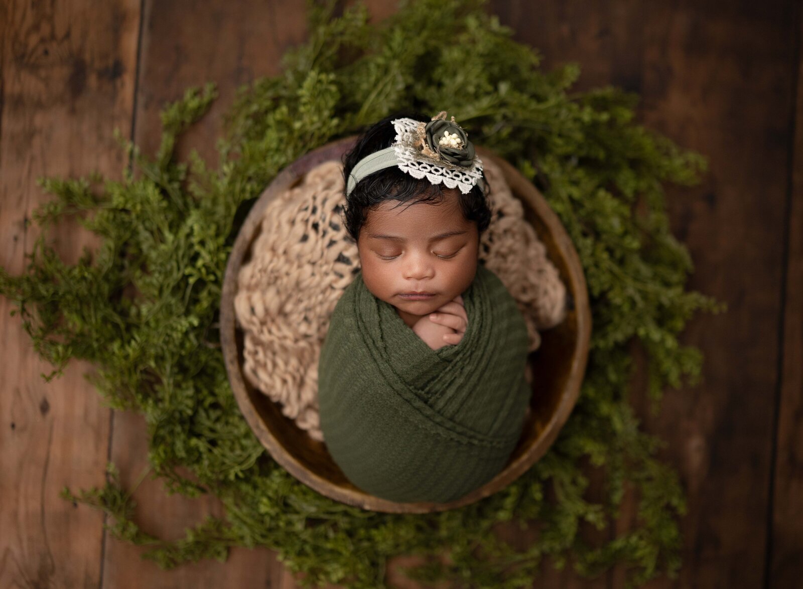 Baby girl swaddled in olive green wrap, laying in a brown bowl with green garland around bowl. Photographed by Jenni Nikki Photography at Marietta Newborn Photo Studio.