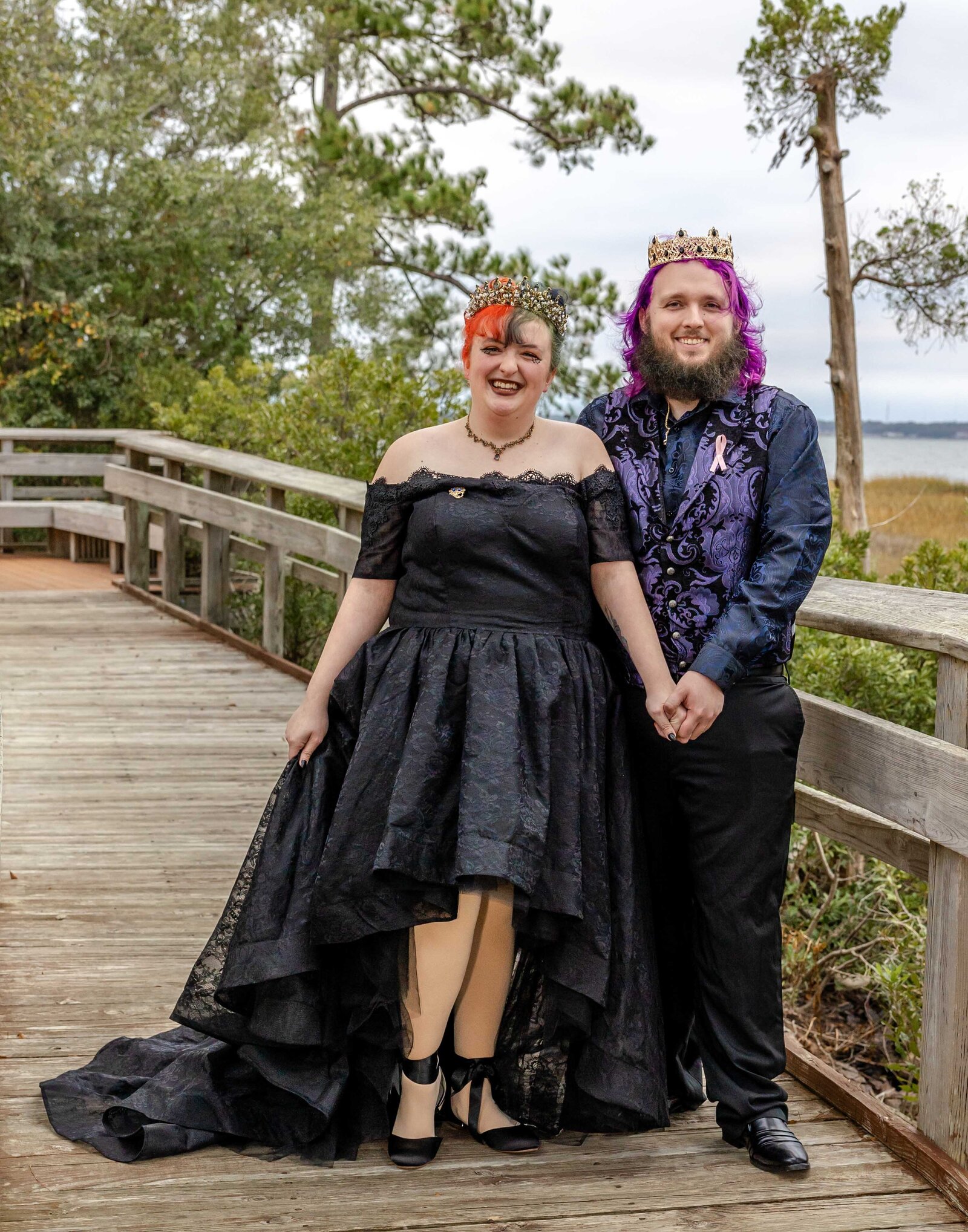 A bride wearing a black wedding dress and a groom in a crown and purple vest pose on a wooden boardwalk surrounded by nature, captured during a unique and creative Pine Knoll Shores, NC Halloween wedding.