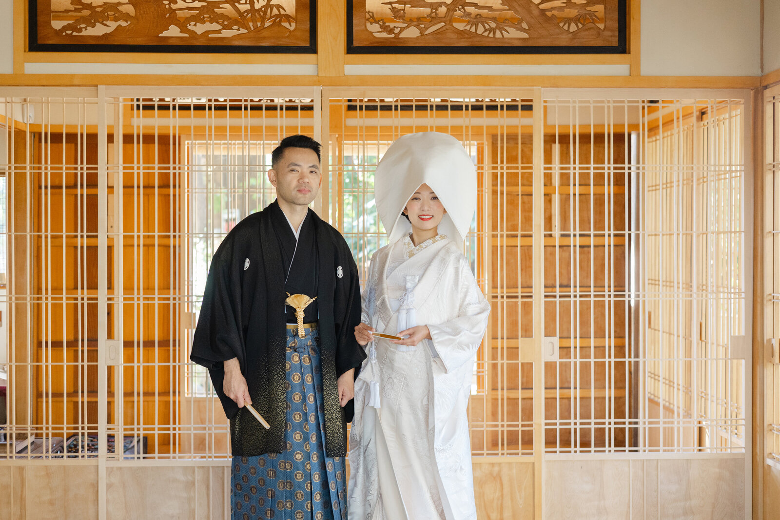 Bride and groom in traditional Japanese wedding attire, standing indoors at the Storrier Stearns Japanese Garden in Pasadena, California. Bride wears a white shiromuku and wataboshi. Available for wedding kimono rental in Los Angeles | カリフォルニア州パサデナのストーリア・スターンズ日本庭園の室内で並ぶ白無垢と綿帽子の花嫁と新郎。着物レンタル可能。