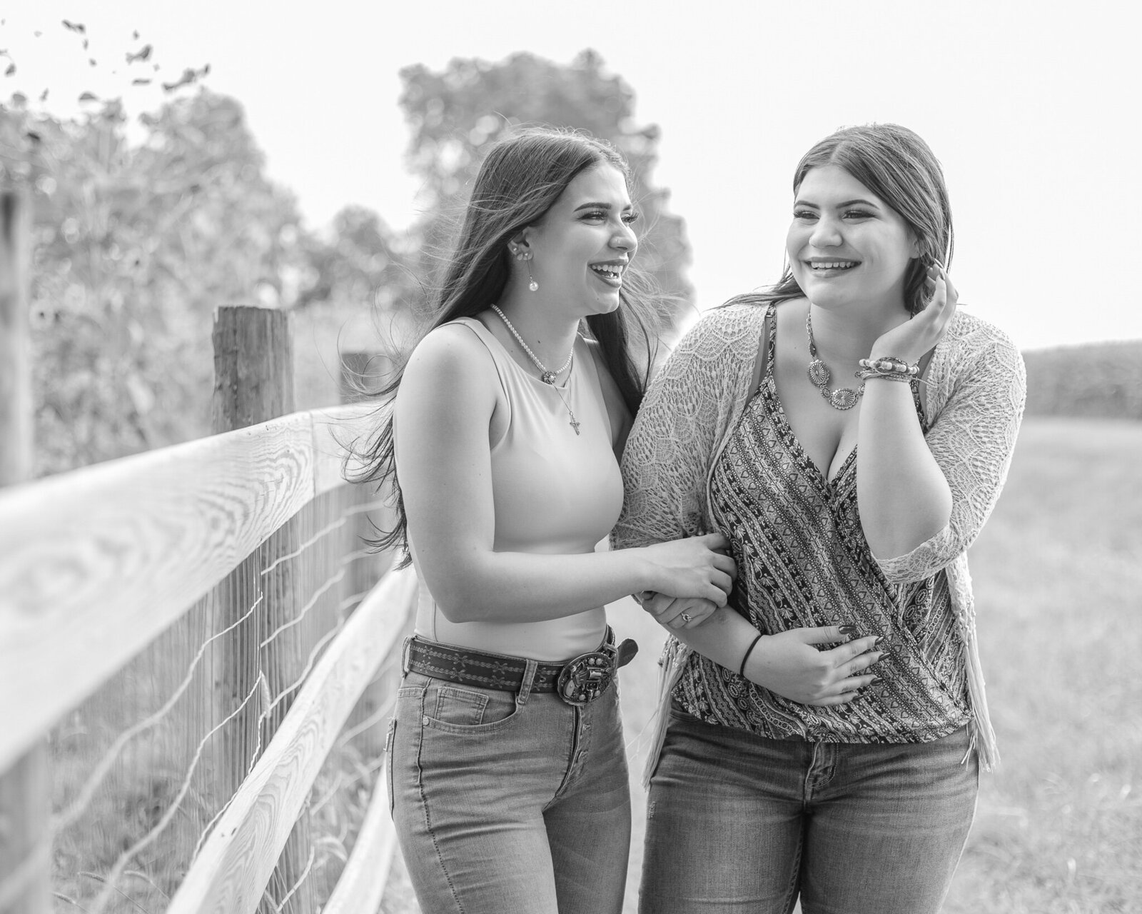 black and white photo of two young ladies, next to a fence that has sunflowers, one of the women has long hair, wearing a sleeveless shirt, belt, and blue jeans, looking at the other woman smiling.  The second woman has her hair down, wearing blue jeans, a patterned shirt, a knit smock over the shirt, tucking her hair behind her ear laughing away from the camera