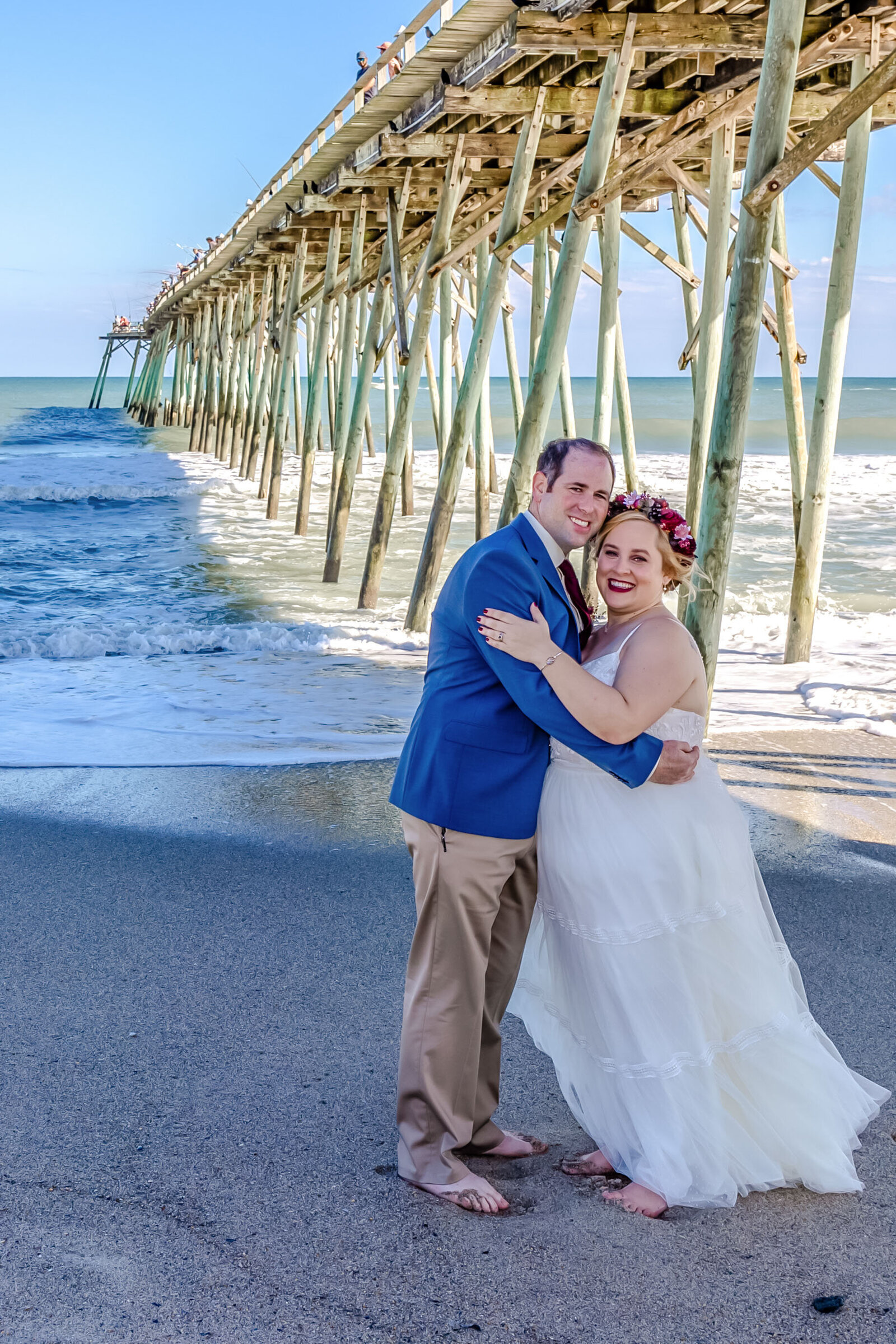 The bride and groom pose together beneath the wooden pier with the ocean waves gently rolling in, captured during a Carolina Beach, NC wedding, blending coastal beauty with romantic charm.