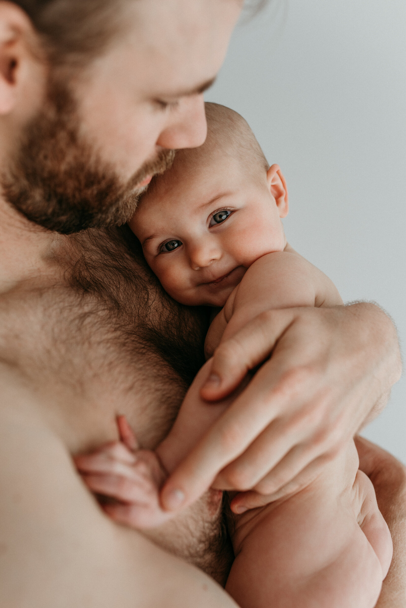 shirtless father holds baby girl while she looks at the camera with a gentle smile and deep eyes