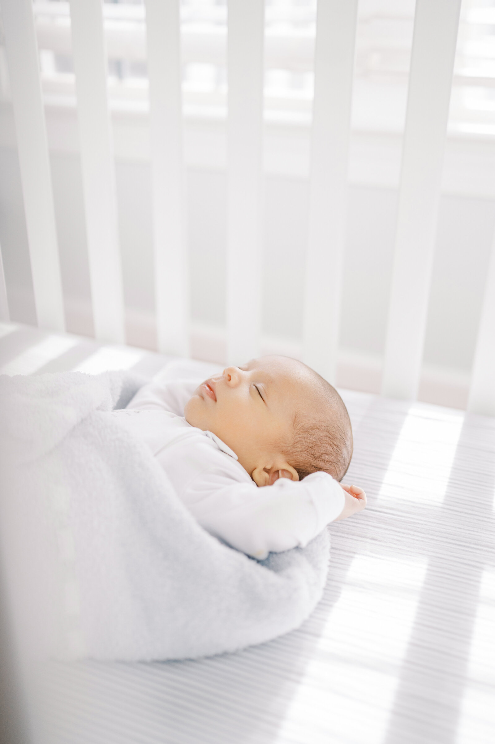 Baby boy wrapped in fuzzy blanket sleeps in white crib in front of a bright window  during in-home newborn session in Raleigh NC