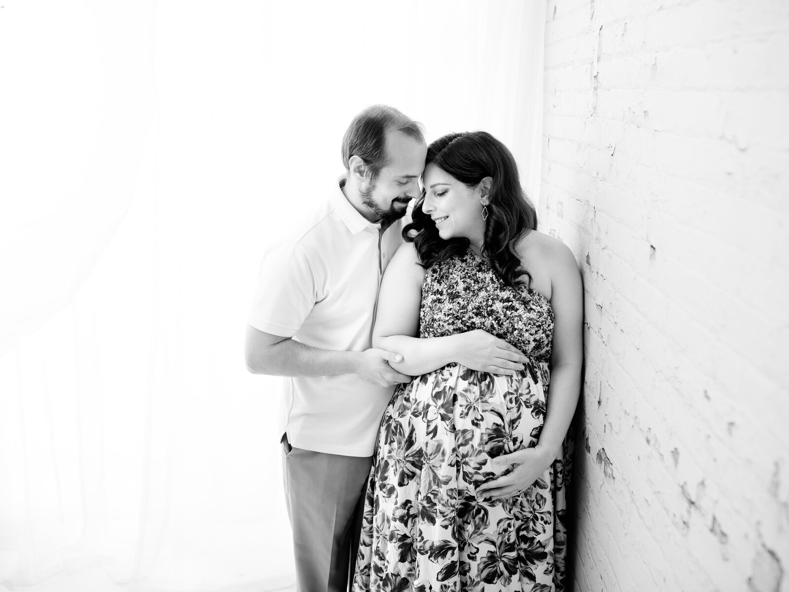 husband and pregnant wife in floral maternity dress holding belly leaning against brick wall