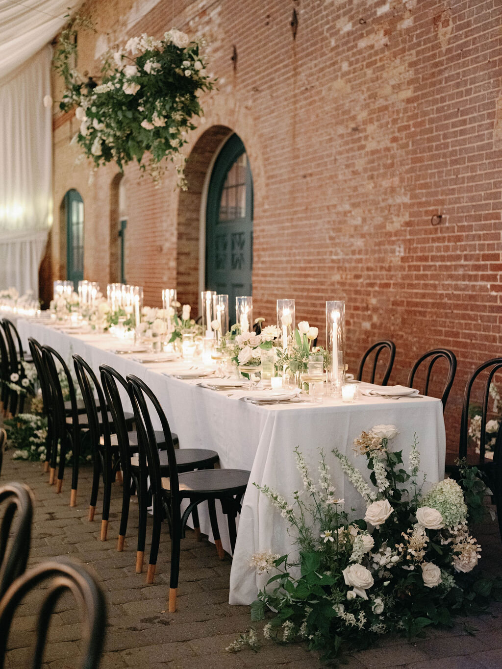 Head table with compote arrangements and pillar candles down the length of the table and ground arrangements at the ends of the table with white larkspur, white roses and white hydrangea Evergreen Museum Carriage House with a hanging installation over the head table
