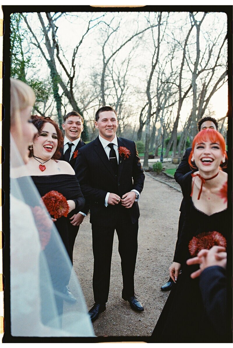 A wedding party stands outdoors, smiling and laughing. The bride, visible from the back, wears a white dress. The others wear black with red accents. Trees and a walking path are in the background. The mood is joyful and celebratory.