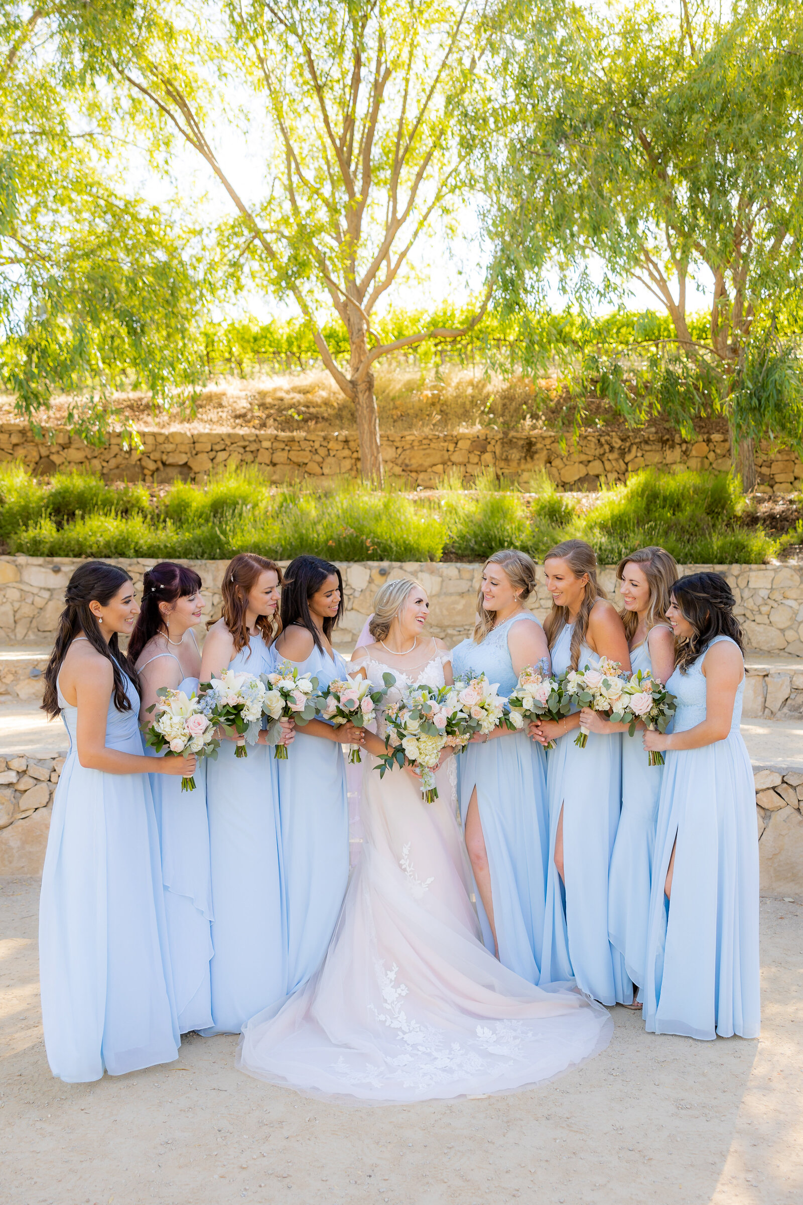 Bridesmaids stand around bride and smiles at her while she smiles back at them. They are wearing a baby blue color while bride is in white.  They are surrounded by greenery on the top. Photo taken by Sacramento Wedding Photographer, Philippe Studio Pro.