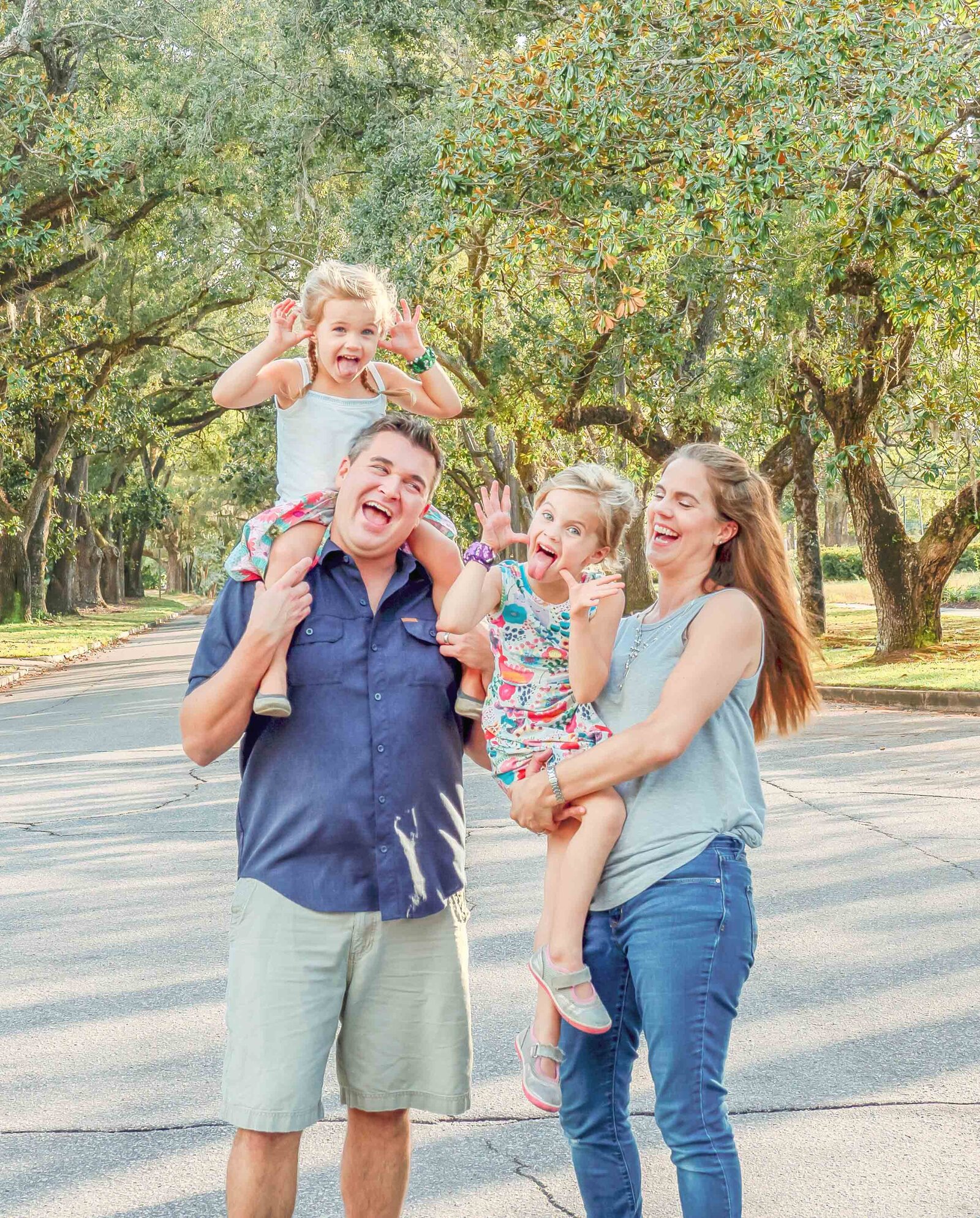 A family shares a playful moment on a tree-lined street, with parents and young children making funny faces. The dappled sunlight creates a warm and inviting atmosphere, perfect for destination family photographer NC, highlighting joyful family connections.