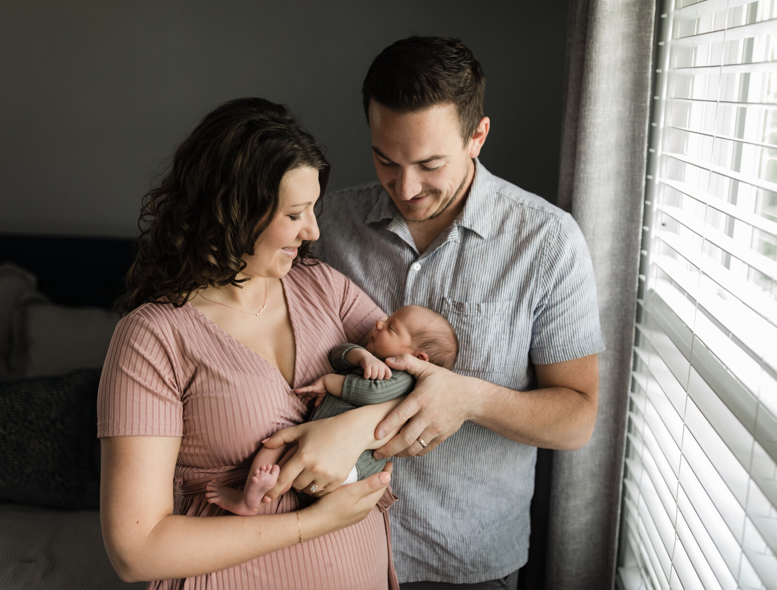mom and dad holding newborn baby boy in bedroom by window