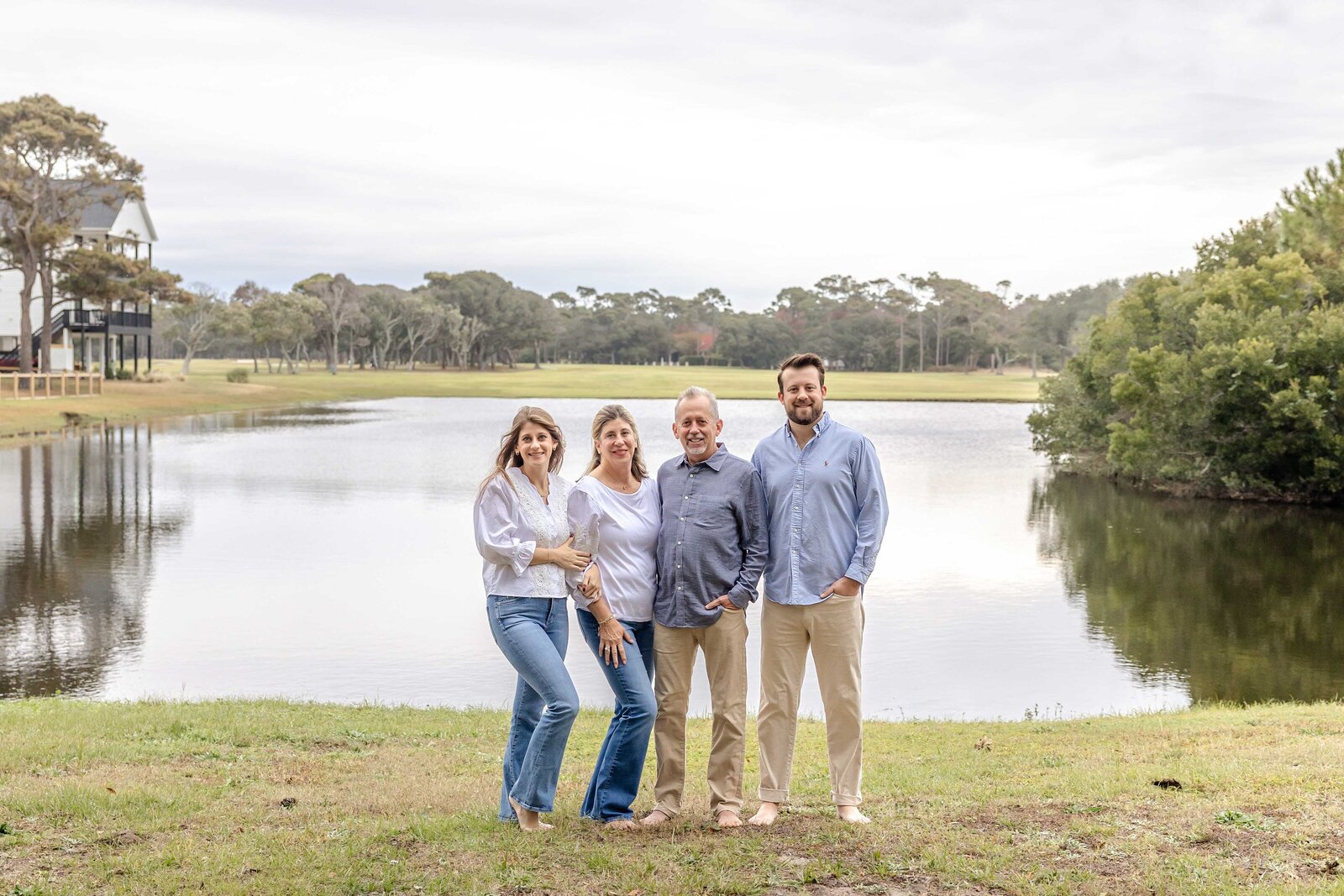 A family of four stands by a serene lakeside, dressed in casual attire. The tranquil water and surrounding greenery create a peaceful backdrop, ideal for North Carolina family photography, capturing relaxed outdoor moments.