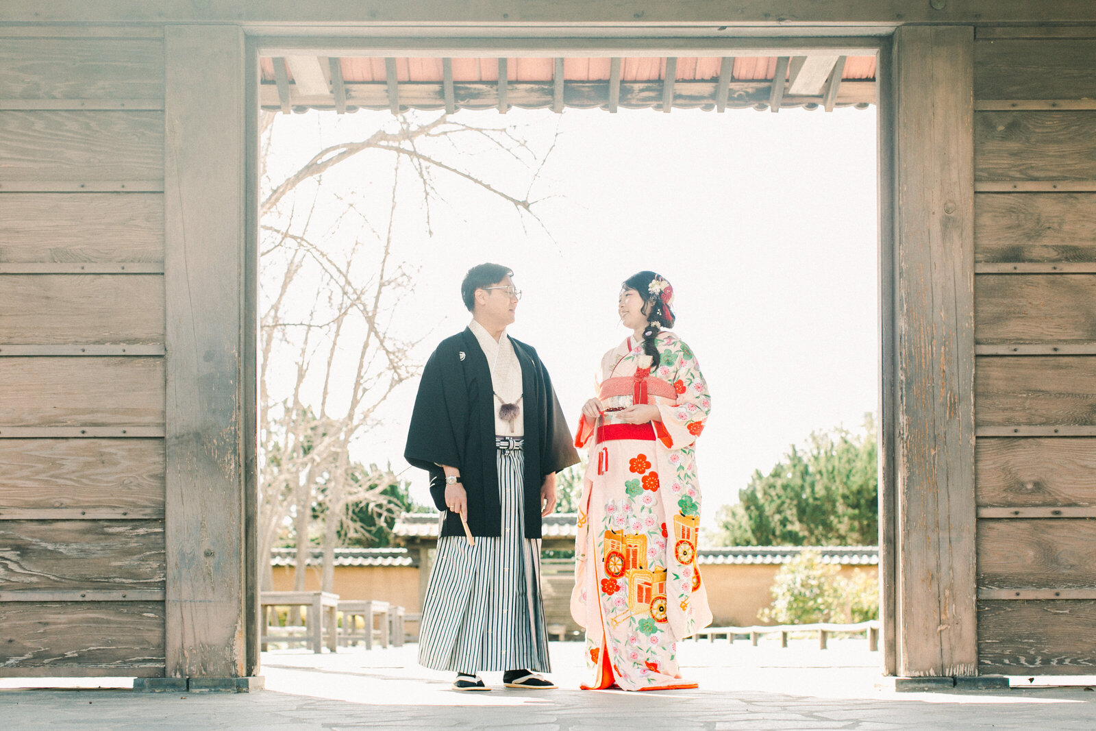 Bride in elegant pink wedding kimono rental walking with groom under the wooden gate at The Huntington Library Japanese Garden, Los Angeles California | ハンティントン図書館日本庭園 木門下での着物レンタルウェディング撮影 新郎新婦