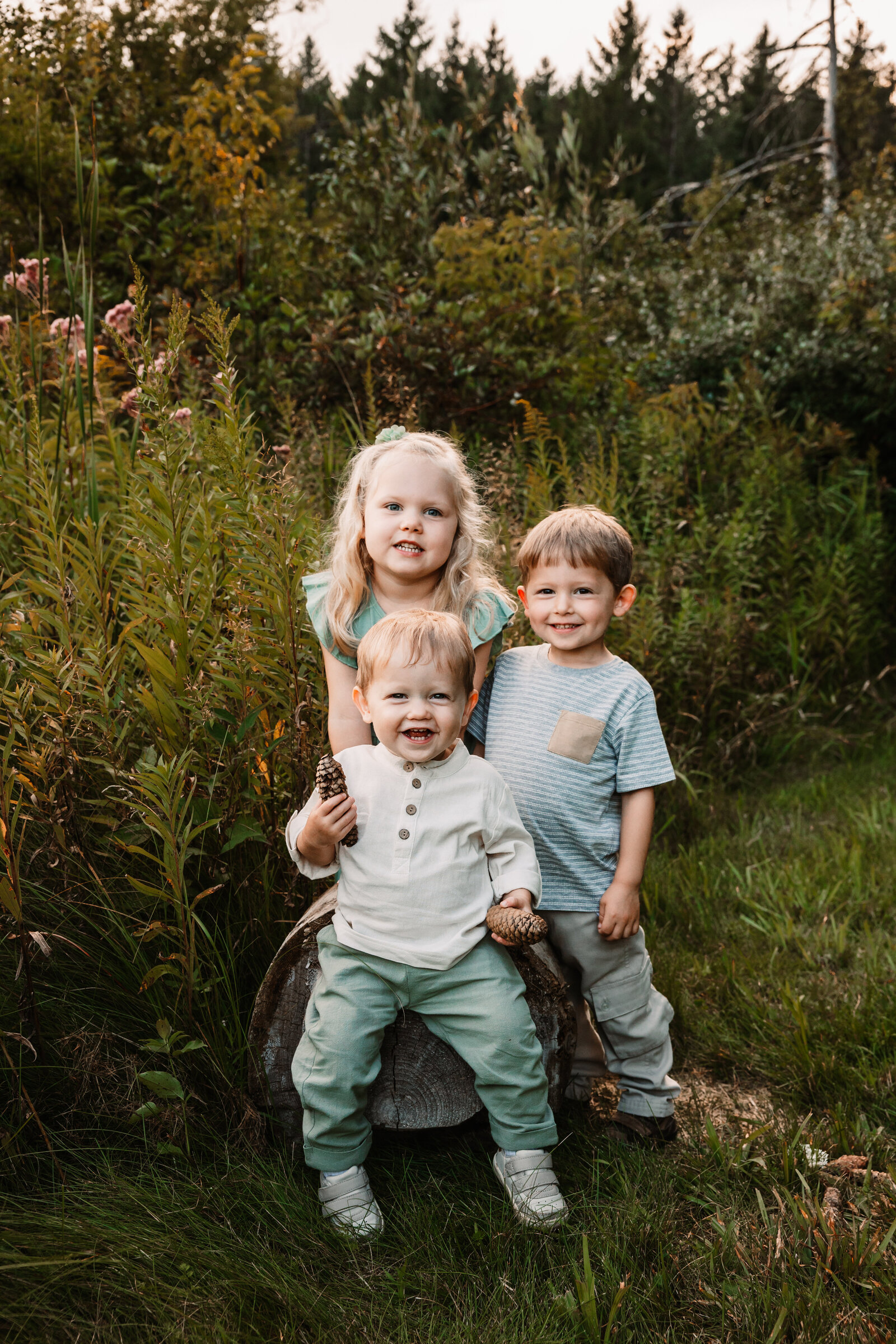 Three toddler siblings laugh and play together on a fallen log in a park