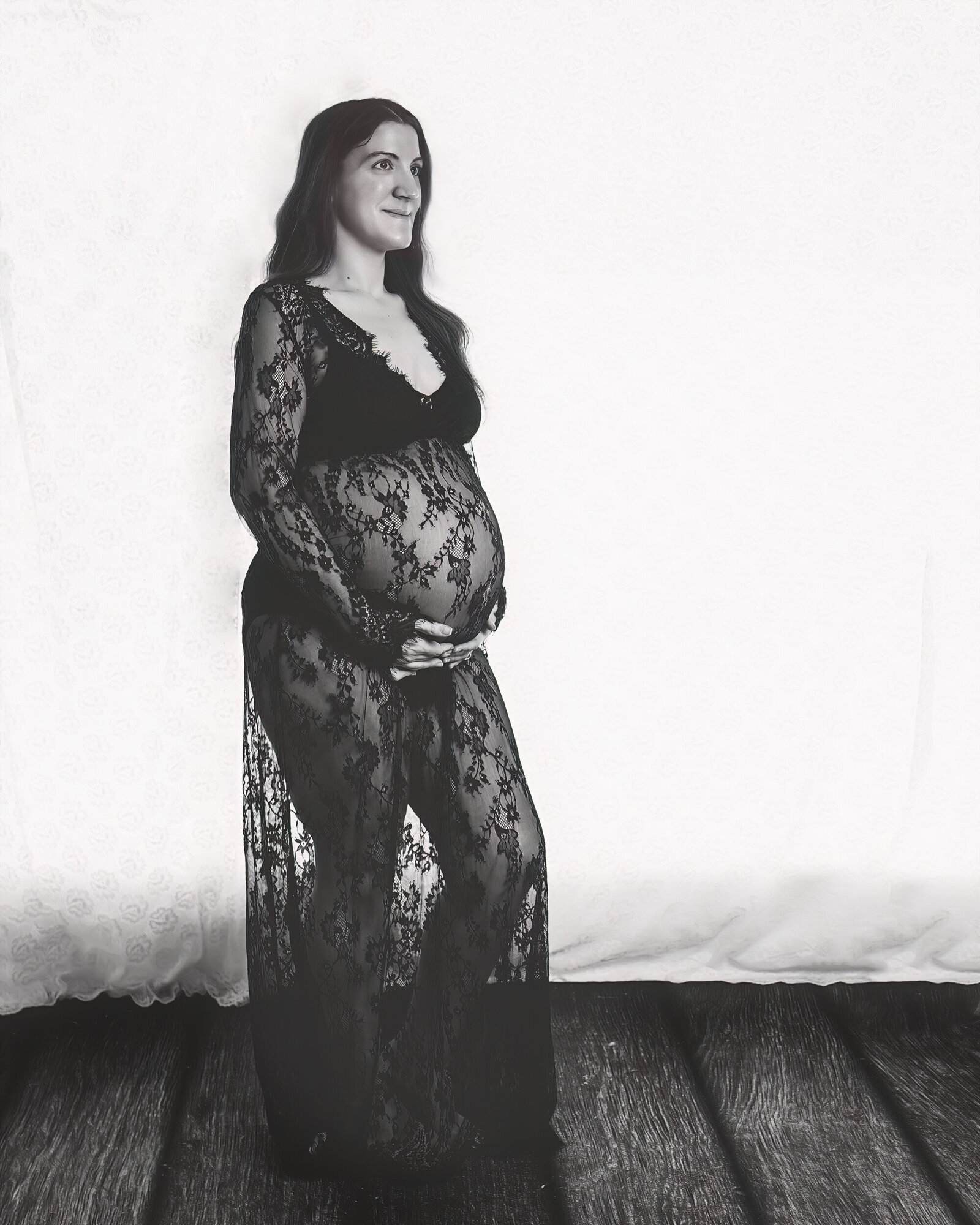 A pregnant woman in a black lace gown cradles her belly while standing against a softly textured white backdrop, exuding timeless elegance.