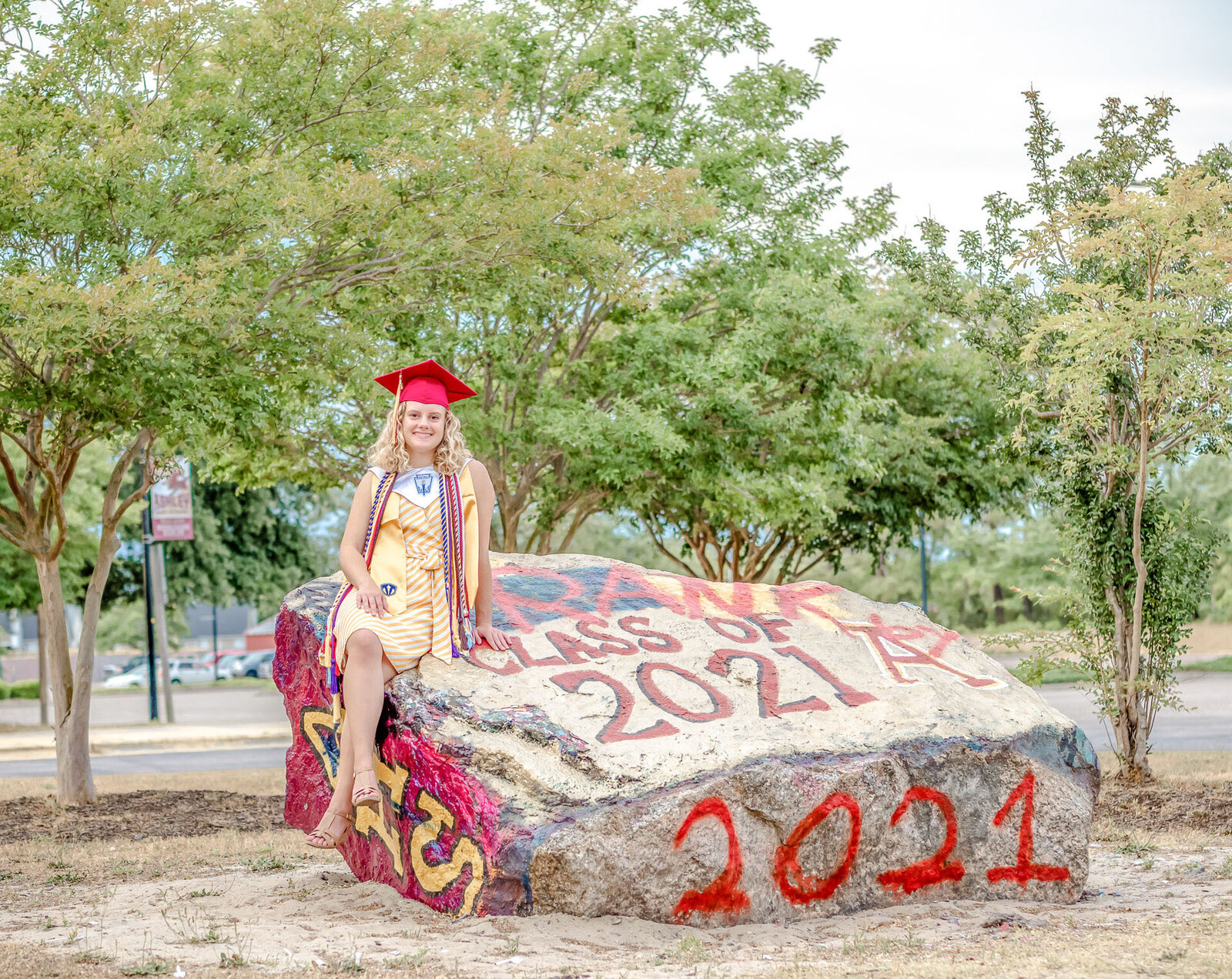 An Ashley High School senior in a red cap sitting on a rock painted with "Class of 2021" in Wilmington, North Carolina. This creative photo captures the celebratory spirit of graduation, ideal for unique senior photography.