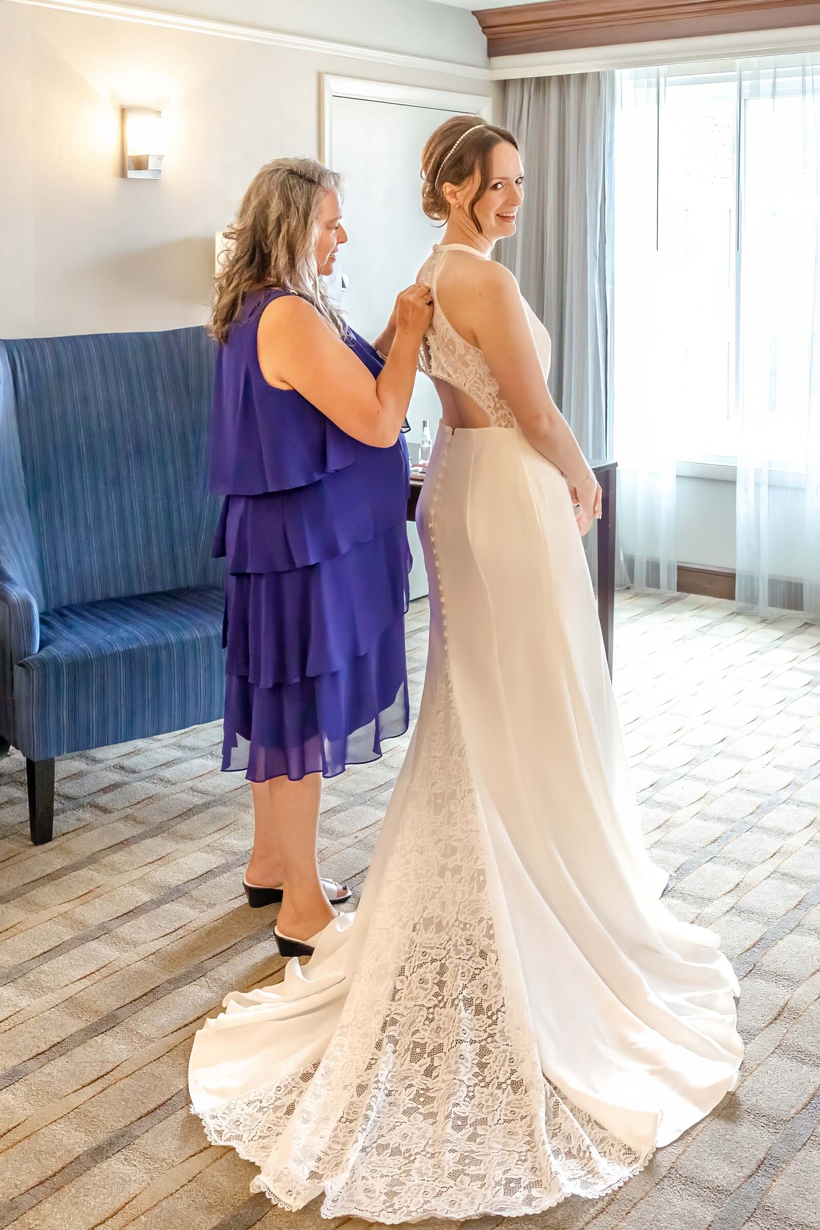 A bride smiles as her mother fastens the back of her lace wedding gown in a bright bridal suite, captured during a Wrightsville Beach, NC wedding, showcasing authentic preparation moments and timeless elegance.