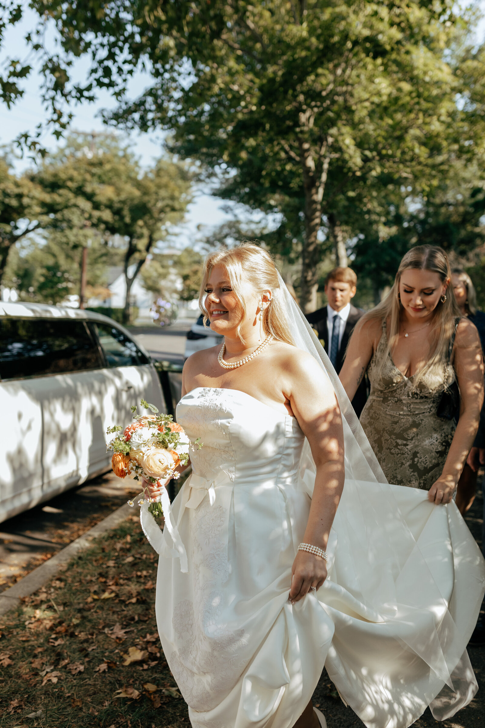Bride walking to her venue with a bridesmaid wearing gold and holding the train of her gown outside during a gorgeous day | Emma LeeAnn Photography