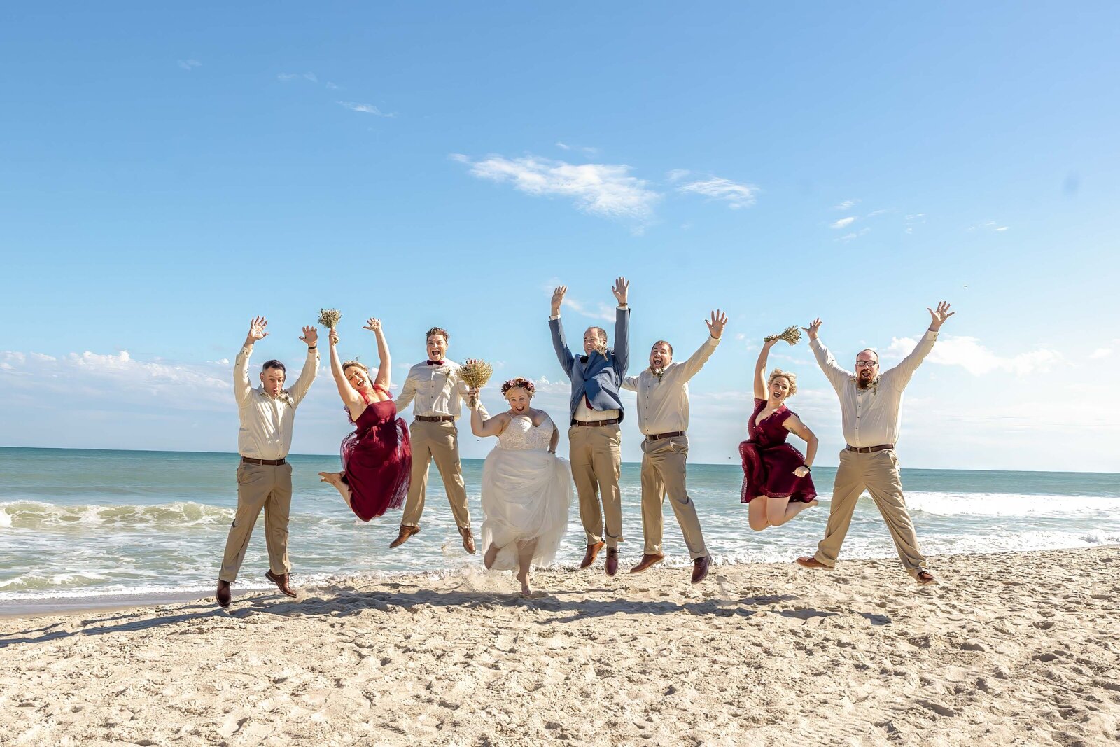 A joyful wedding party jumps in the air on the sandy shores of Topsail Beach, NC, with the ocean and clear blue sky in the background, showcasing fun and authentic moments at a coastal wedding.