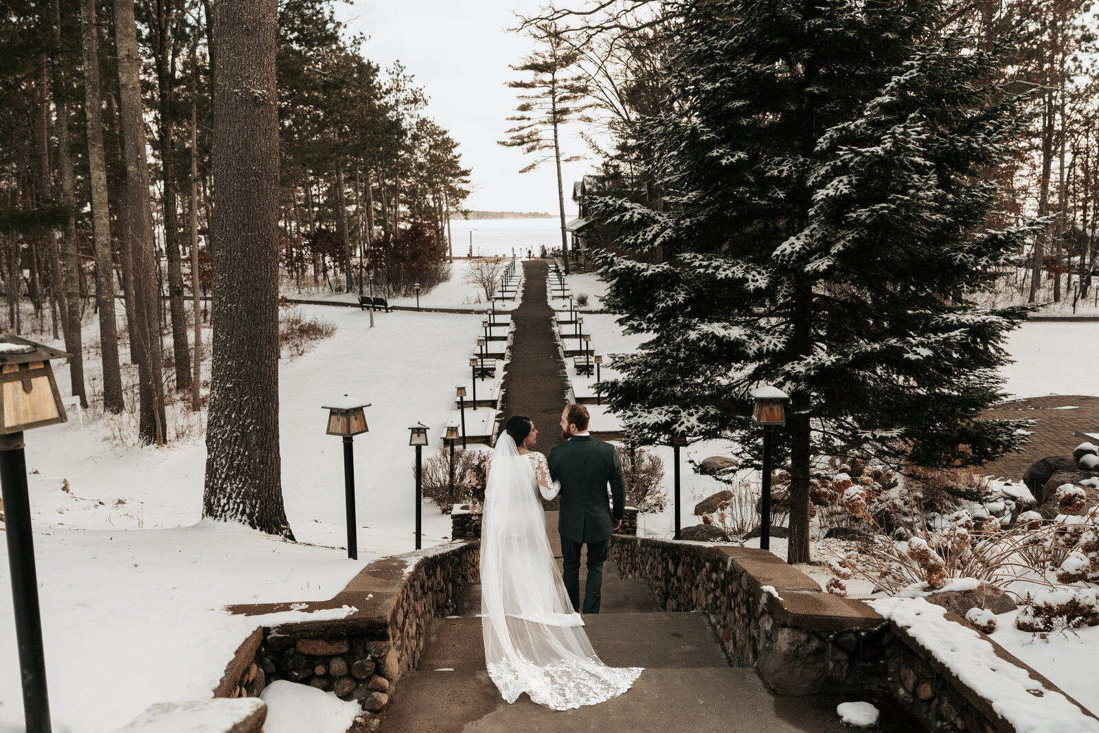 Couple stands on a snow-covered stone path overlooking a winter lake, surrounded by evergreen trees in a scenic Minnesota elopement setting.