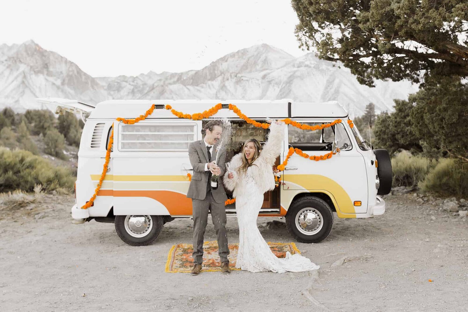 A bride and groom pop champagne in front of their vintage VW kombi, in front of a snow capped Mammoth Lakes mountain. 