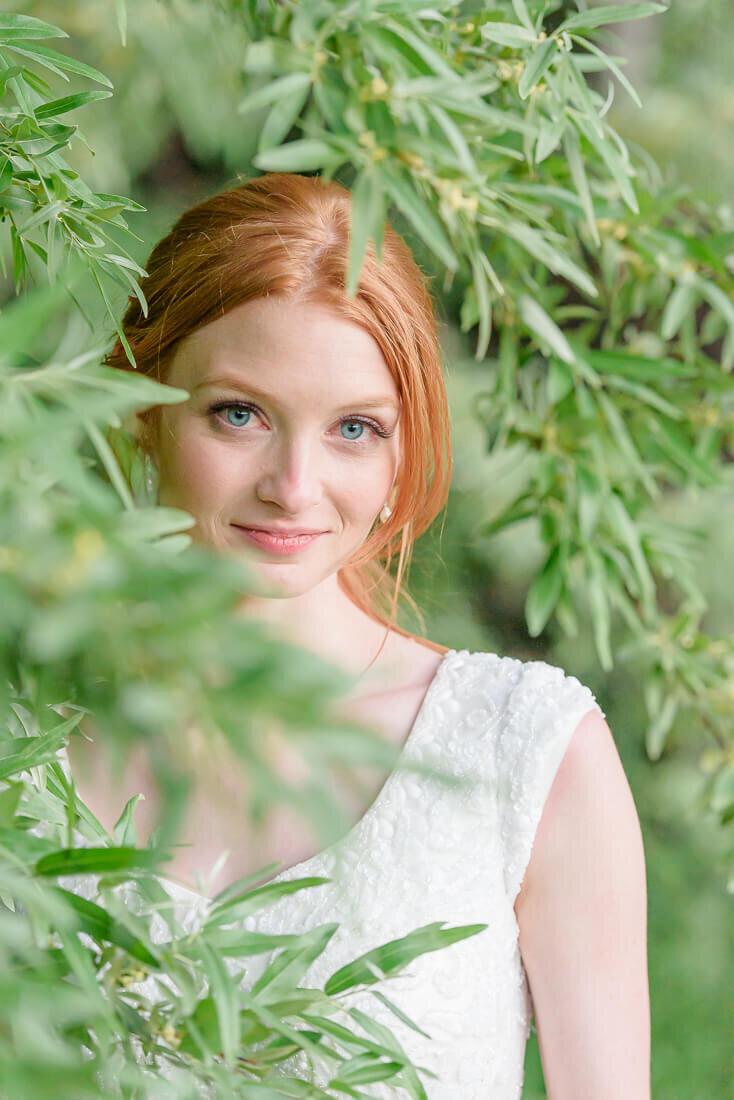 A summer bride with red hair wearing an off white wedding dress covered in beads and sequins stands in green trees. Captured at the River Oaks Golf Course in Sandy