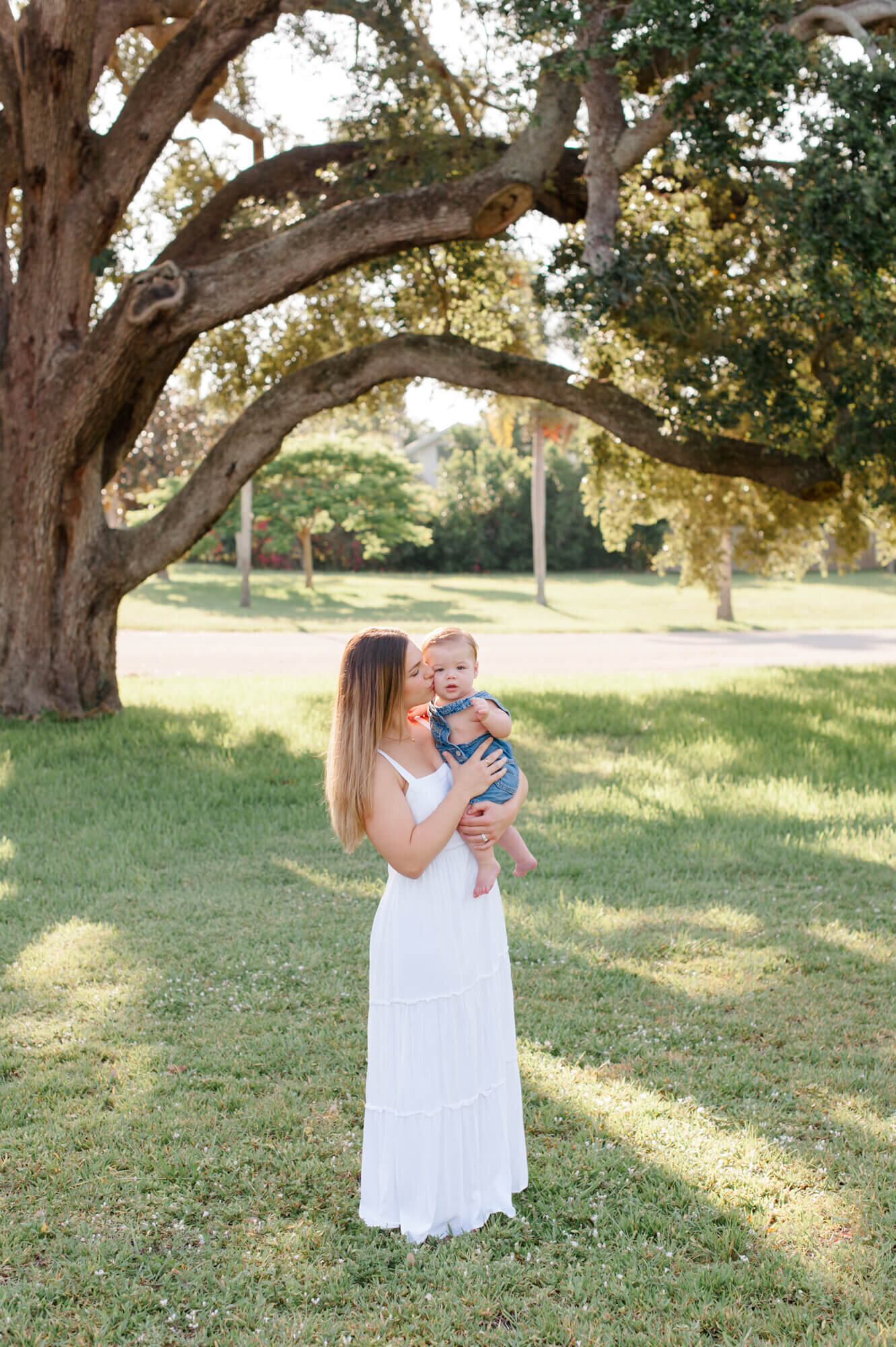 Mom holds her son and kisses his cheek while standing underneath an oak tree