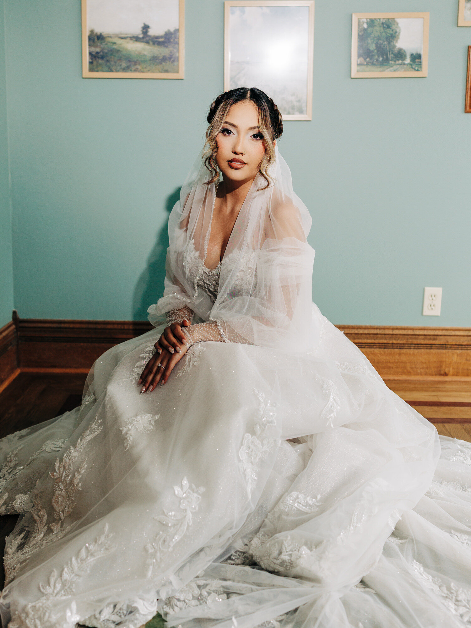 A woman in a white wedding dress with lace details sits on a wooden floor against a teal wall with framed art—captured beautifully by a San Antonio Wedding Photographer. Her hair is styled in curls, makeup natural, her expression calm and poised.