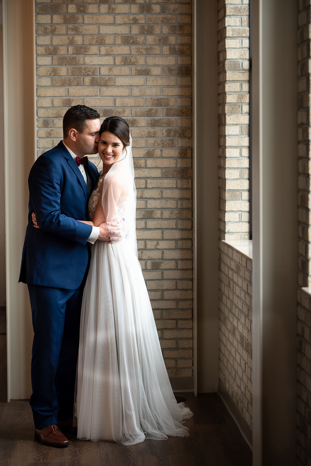 Husband kissing a wifes cheek  by a window in front of a brick wall