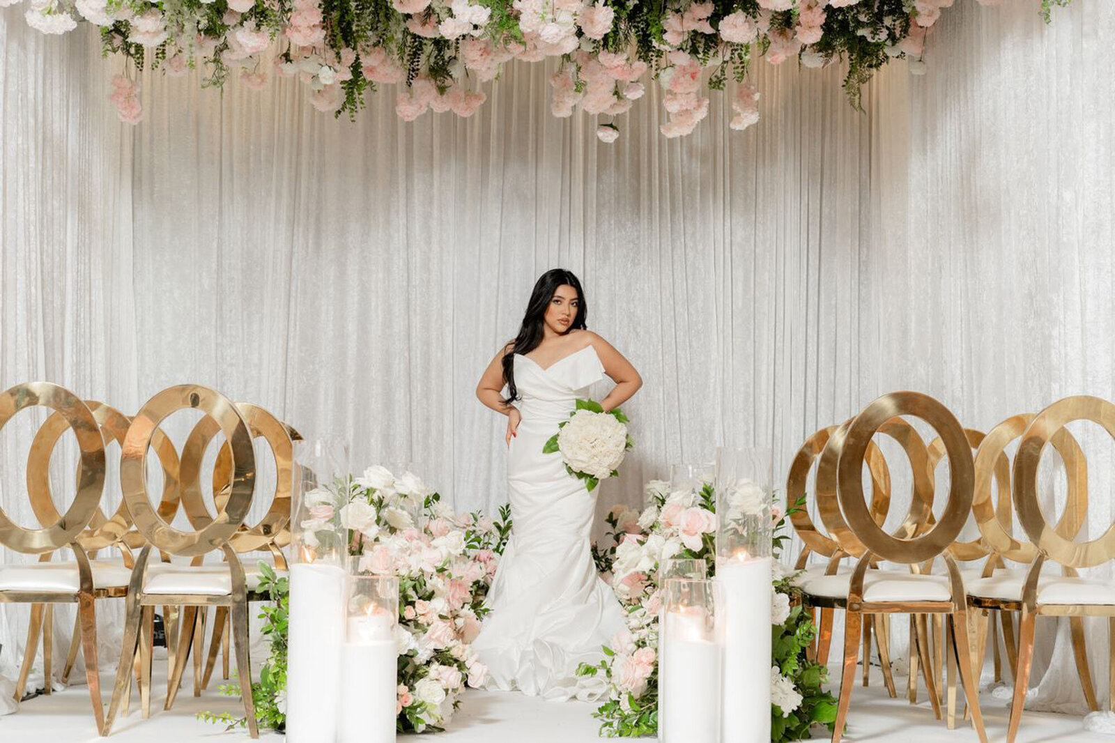 woman wearing wedding dress and holding a white bouquet, standing in front of gold chairs