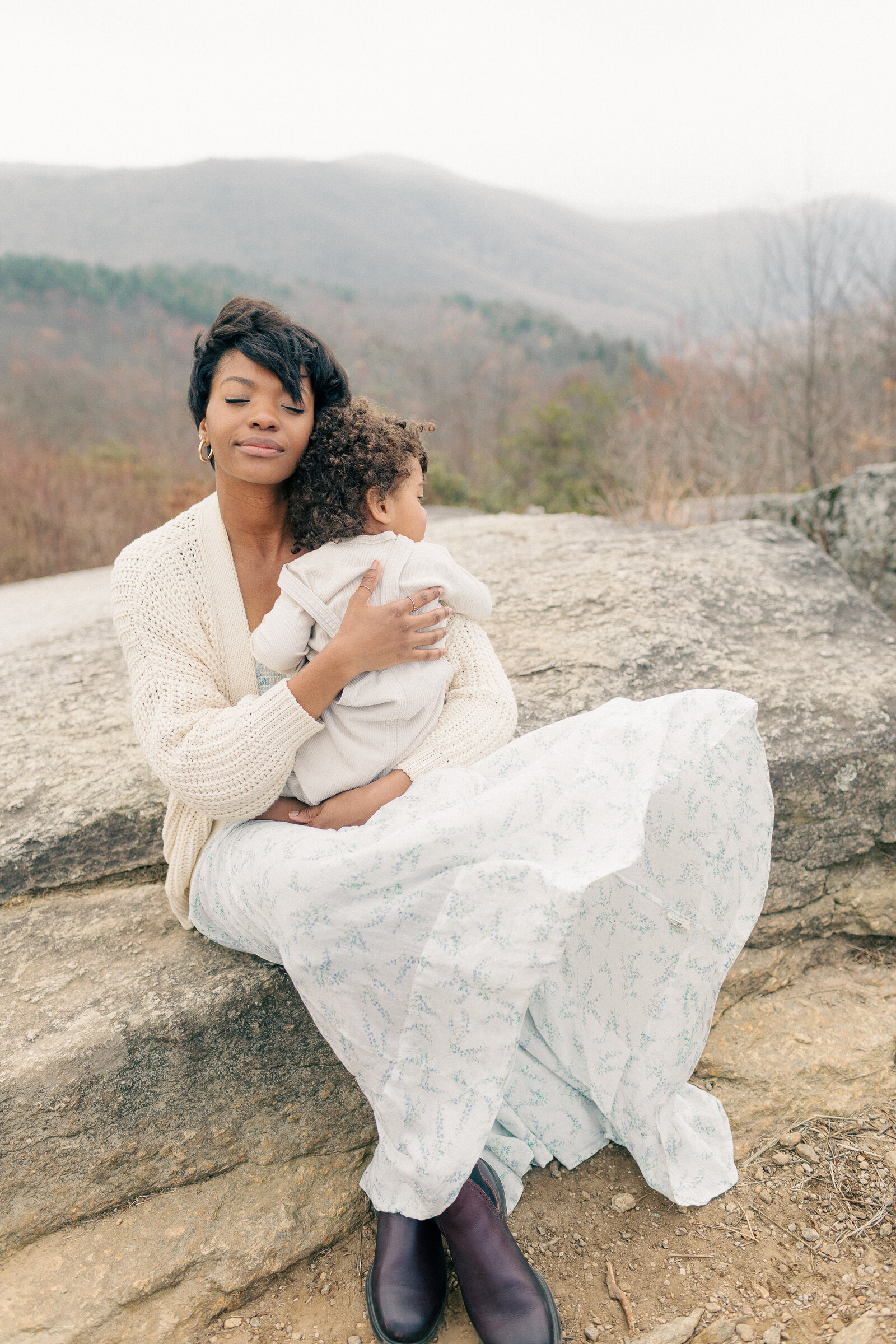 Mother sitting on a large rock holding her toddler in her lap, both dressed in soft neutrals during an outdoor Kansas City family session.