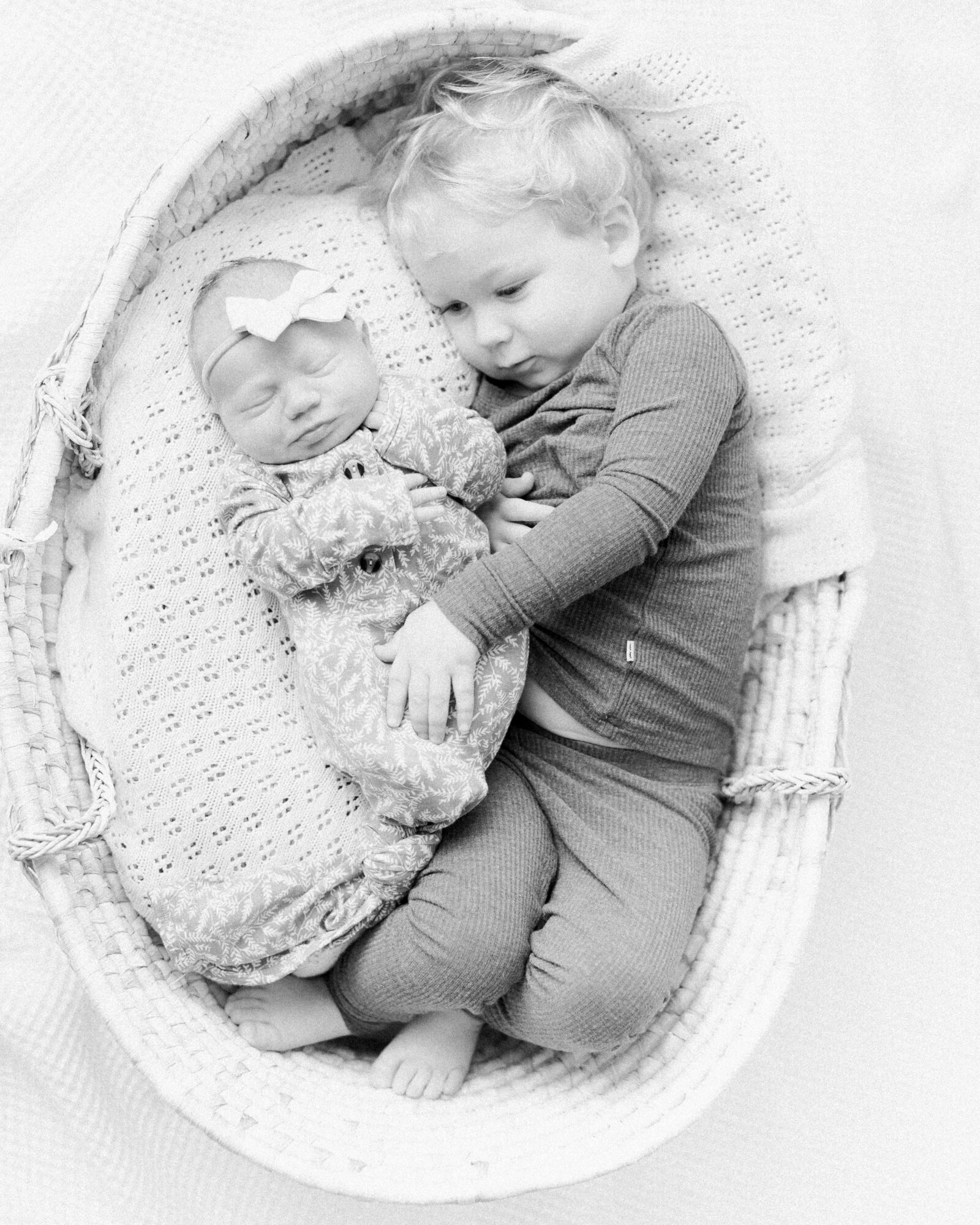 Toddler brother snuggles next to his baby sister with his hand soothing her on a white crochet blanket in a moses basket by NH newborn photographer Fieldstone Studio.