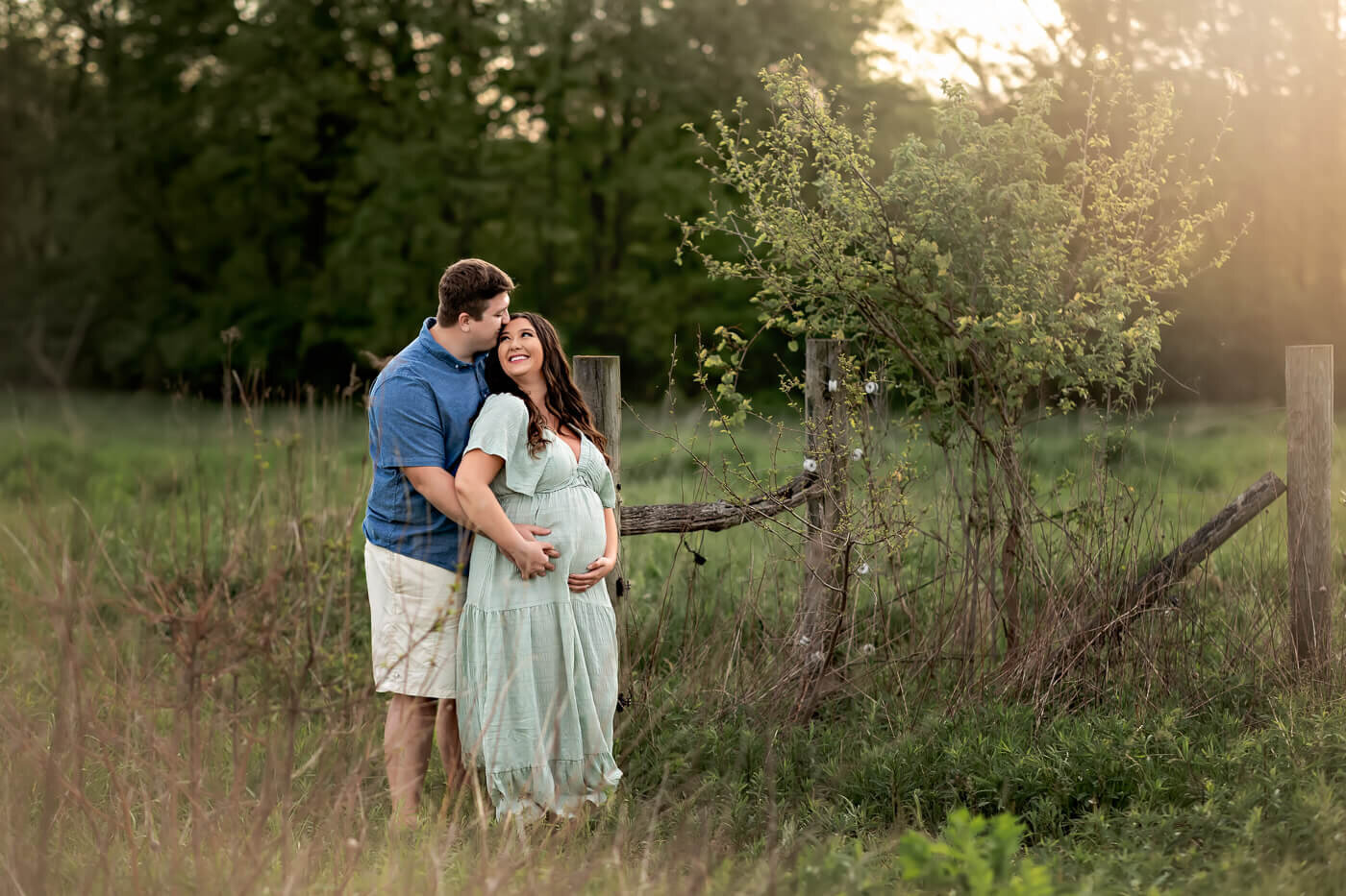 A smiling pregnant woman in a light green dress stands in a grassy field, embraced by her partner near a wooden fence and small tree, bathed in warm sunlight—capturing the joy of maternity portraits with a Bloomington maternity photographer.