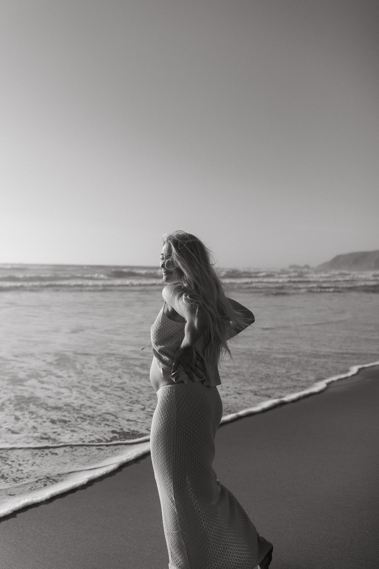 Mother-to-be in flowing dress walking along the shoreline at Point Reyes