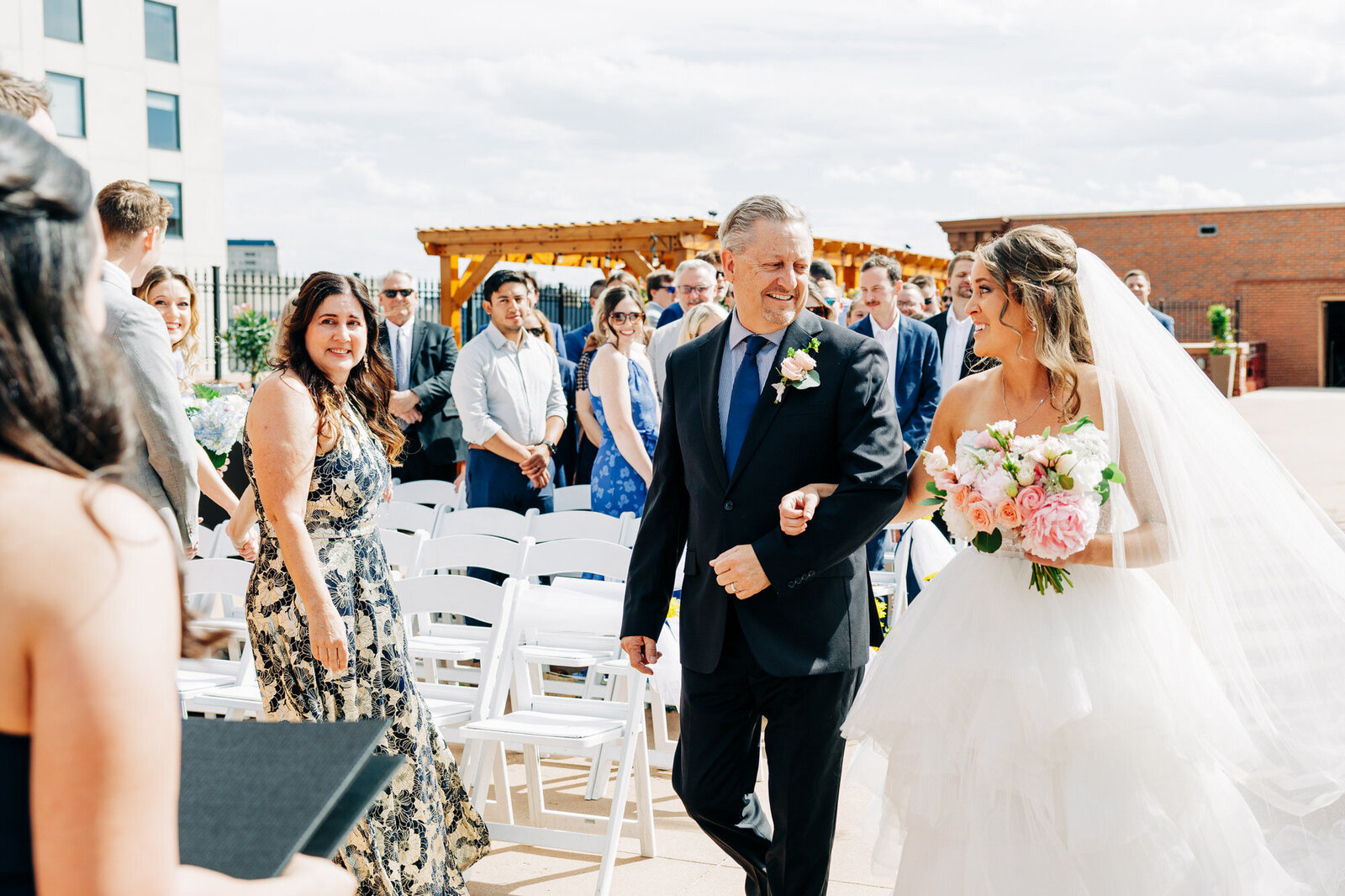 Austin wedding photographer captures a bride in a white dress and veil walks down the aisle with an older man in a suit, both smiling. Guests seated and standing nearby watch happily at an outdoor wedding ceremony. The bride holds a bouquet of pink and white flowers.